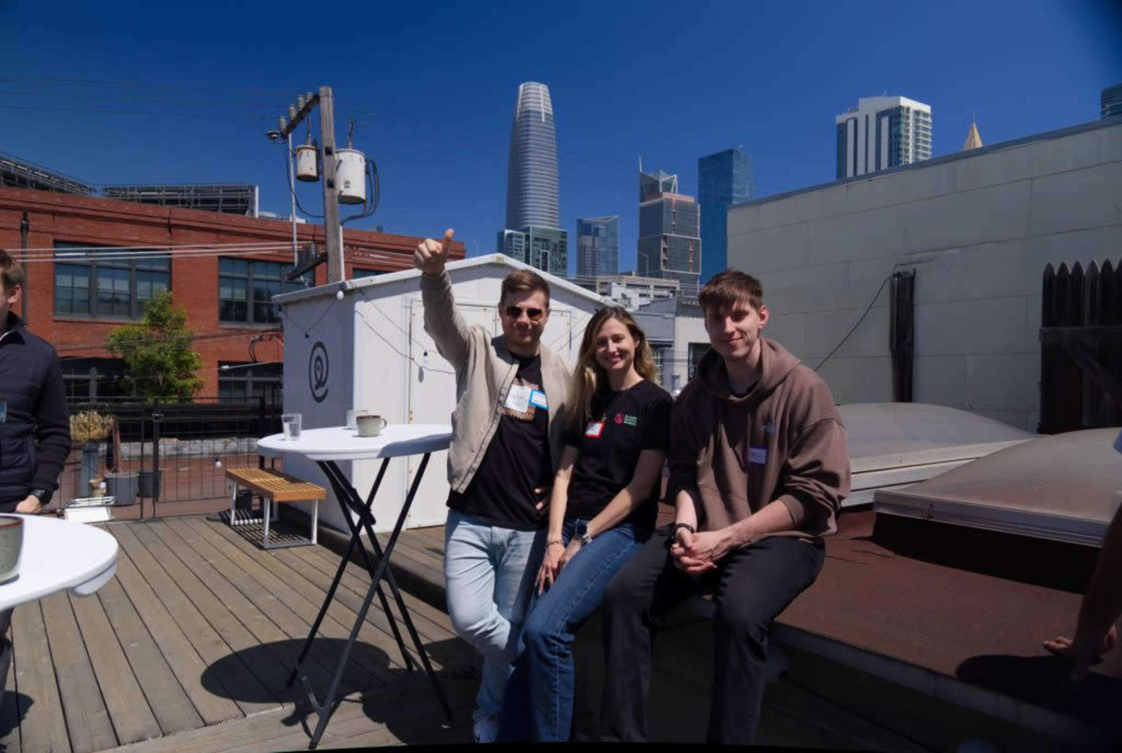Three people pose on a rooftop with a city skyline in the background featuring tall buildings.