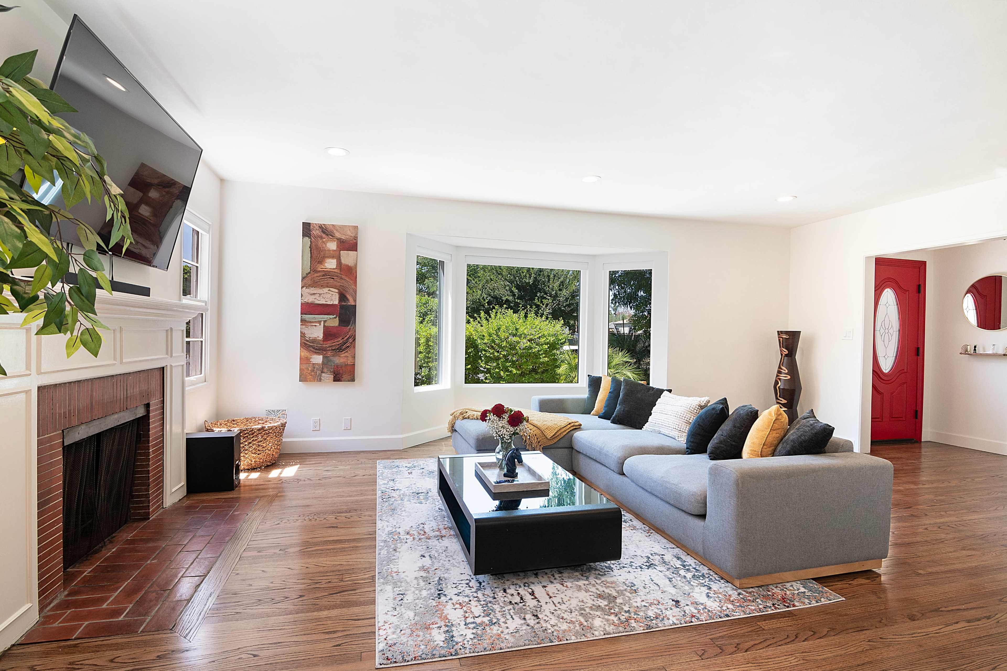 The living room features a gray sofa, a coffee table, and a large window with views of greenery, along with a flat-screen TV mounted on the wall.