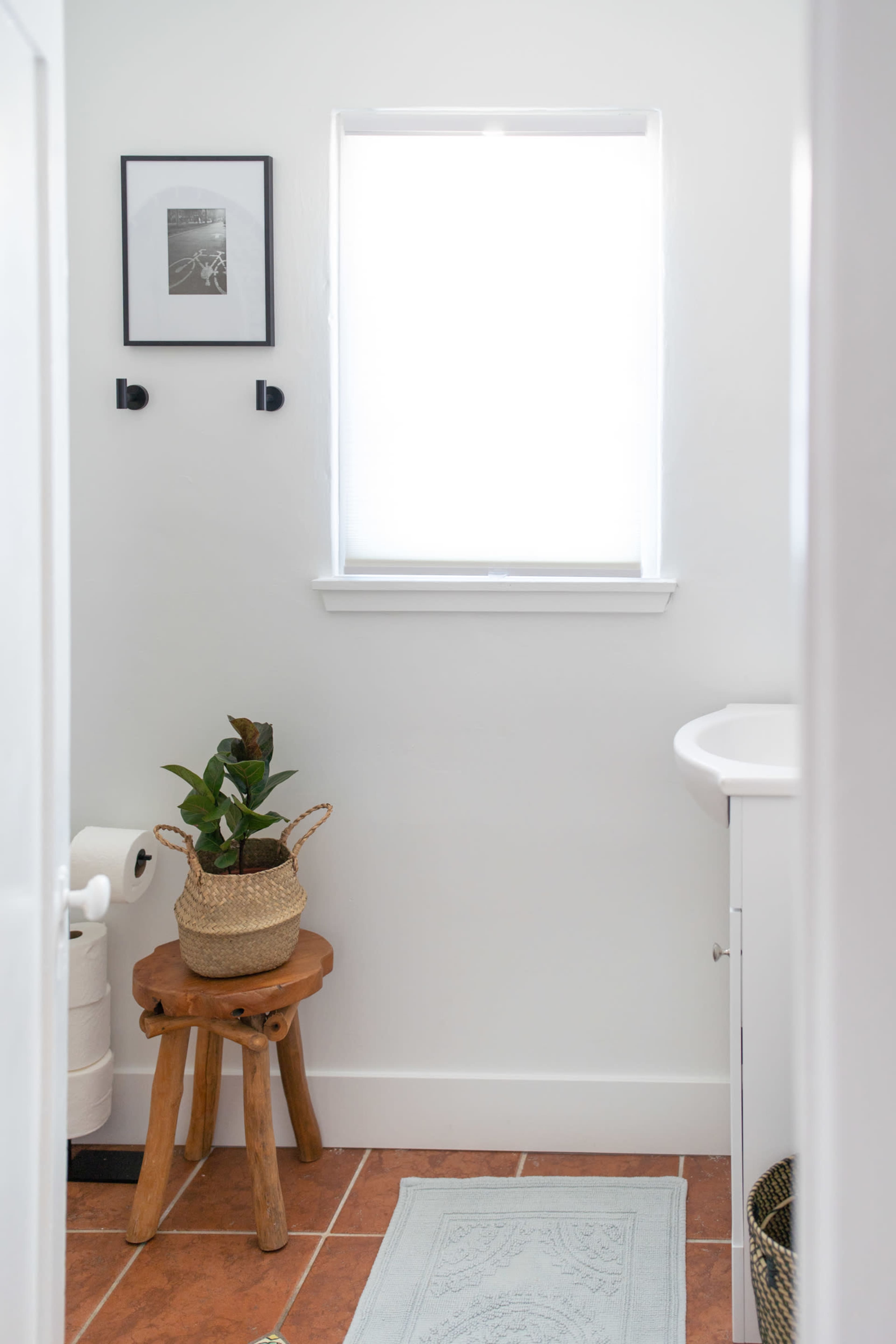 A bright bathroom with a window, a small plant on a wooden stool, and a sink against a white wall.