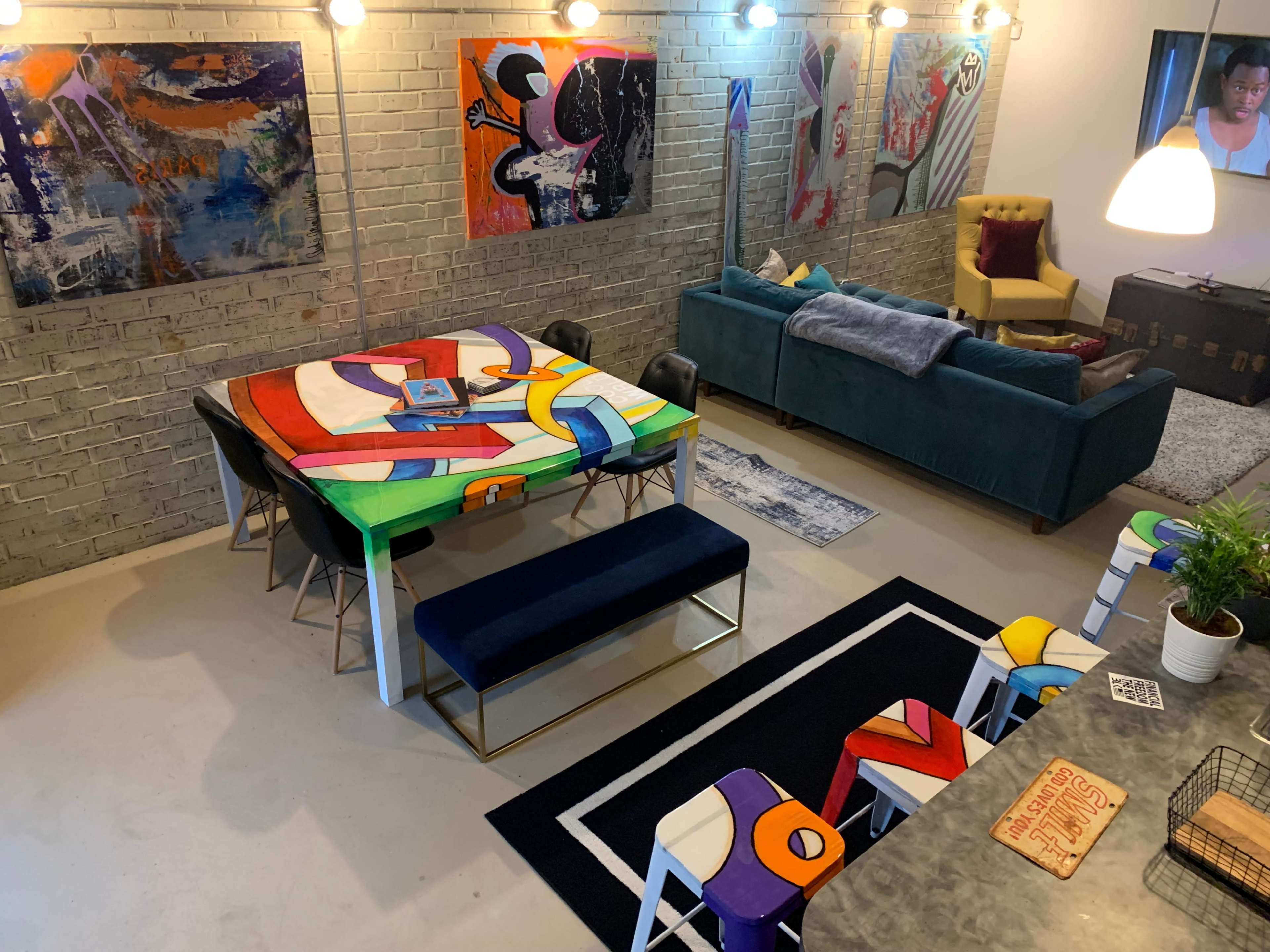 A colorful dining area with a large, artistic table surrounded by sleek chairs, a cozy sofa, and various artworks displayed on the brick walls.
