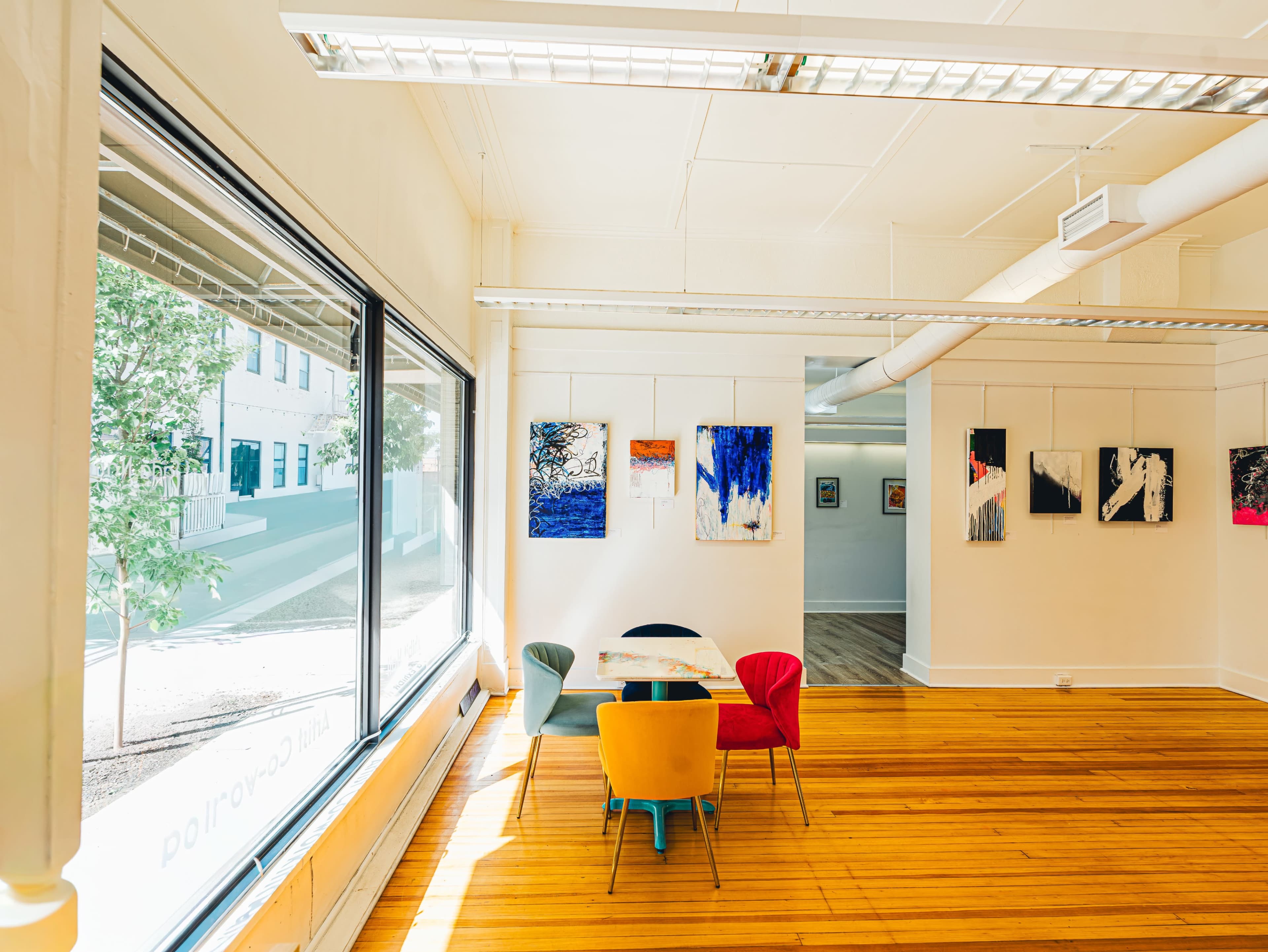The image shows a bright, modern office space featuring a round table surrounded by colorful chairs and an art display on the walls.