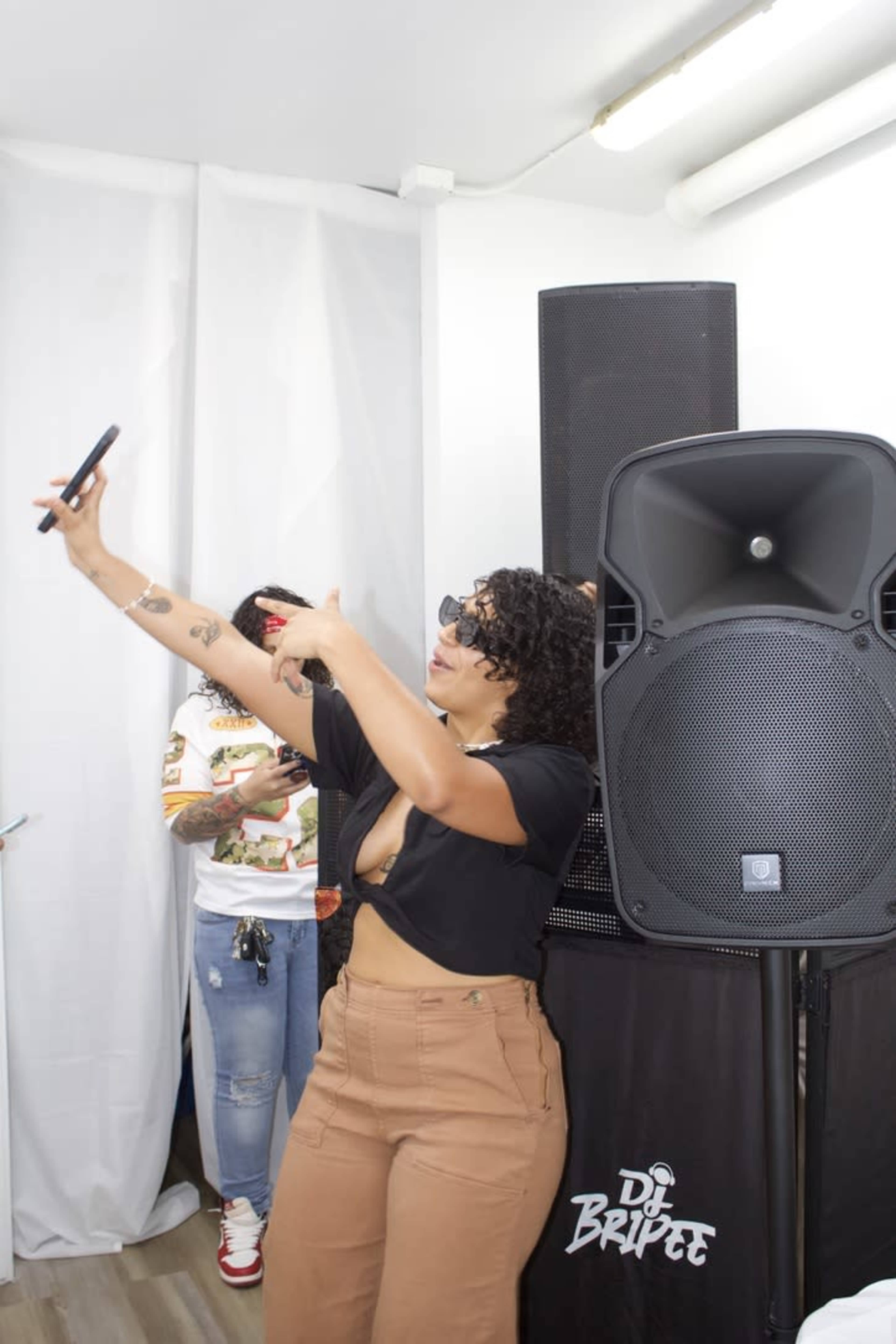 A woman is taking a selfie in front of a large speaker at an event, while another person is visible in the background.