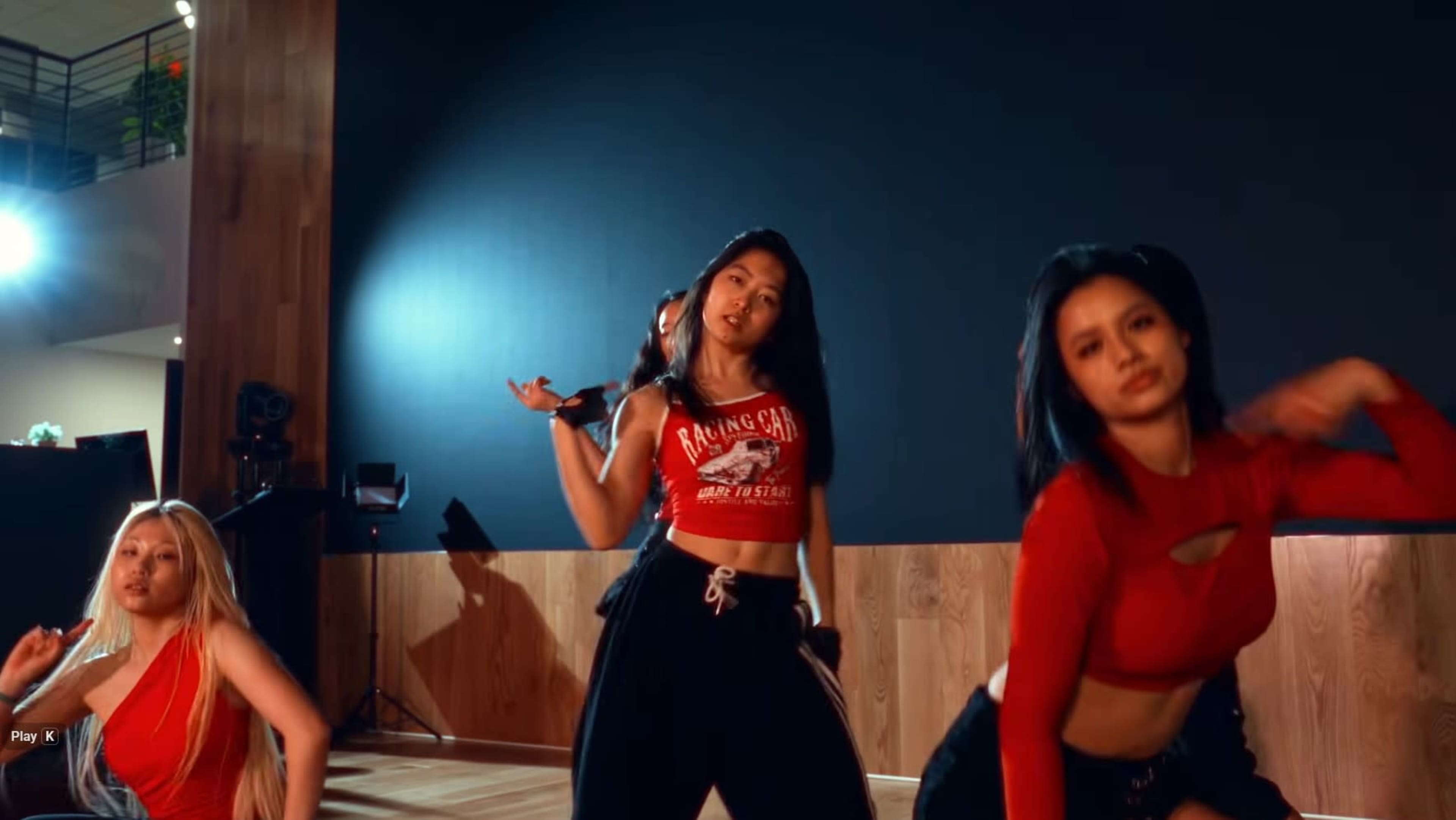 Three dancers perform together in a studio with wooden flooring and a dark wall backdrop.