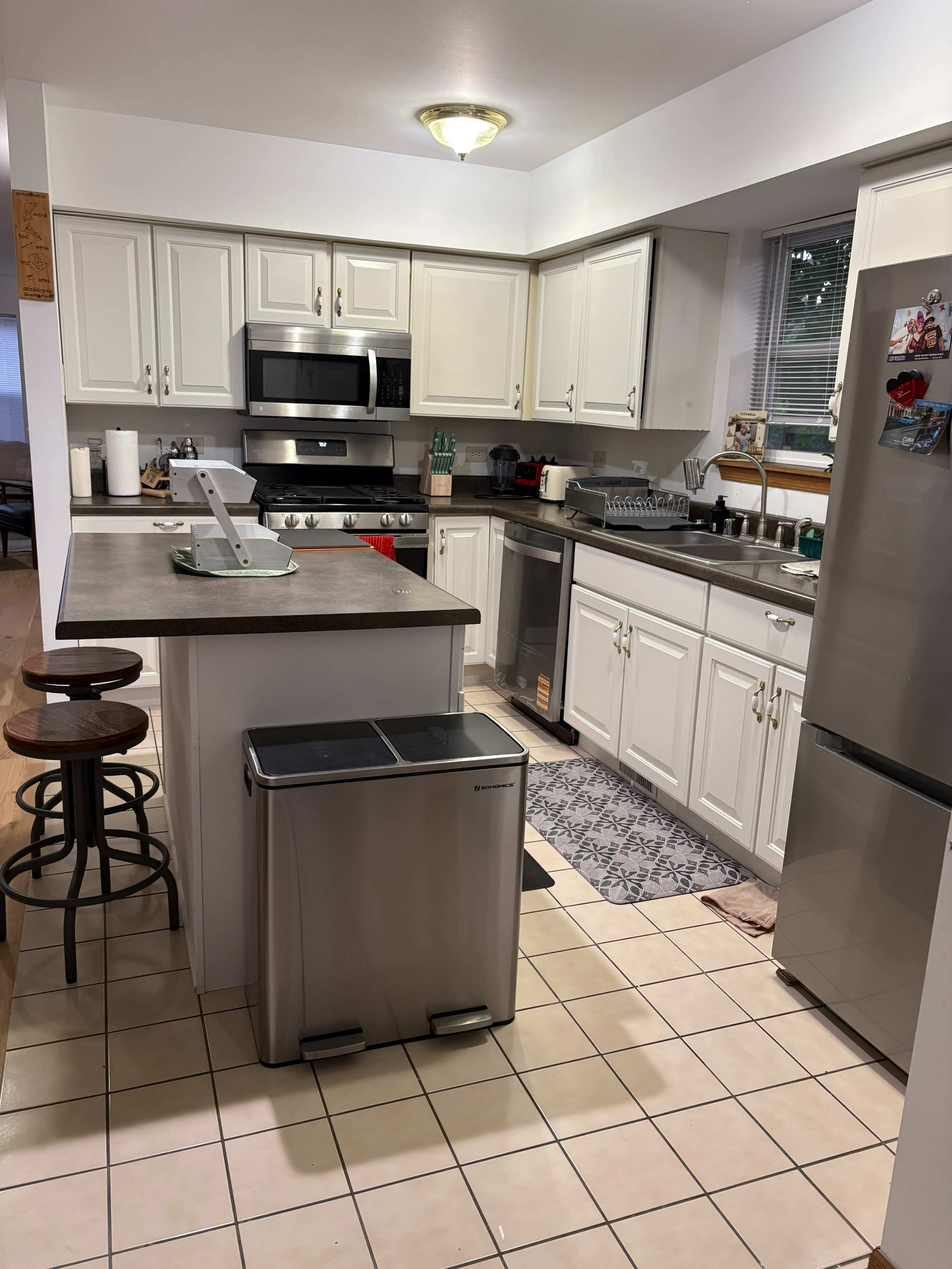 The image shows a kitchen with white cabinets, stainless steel appliances, and a central island with barstools.