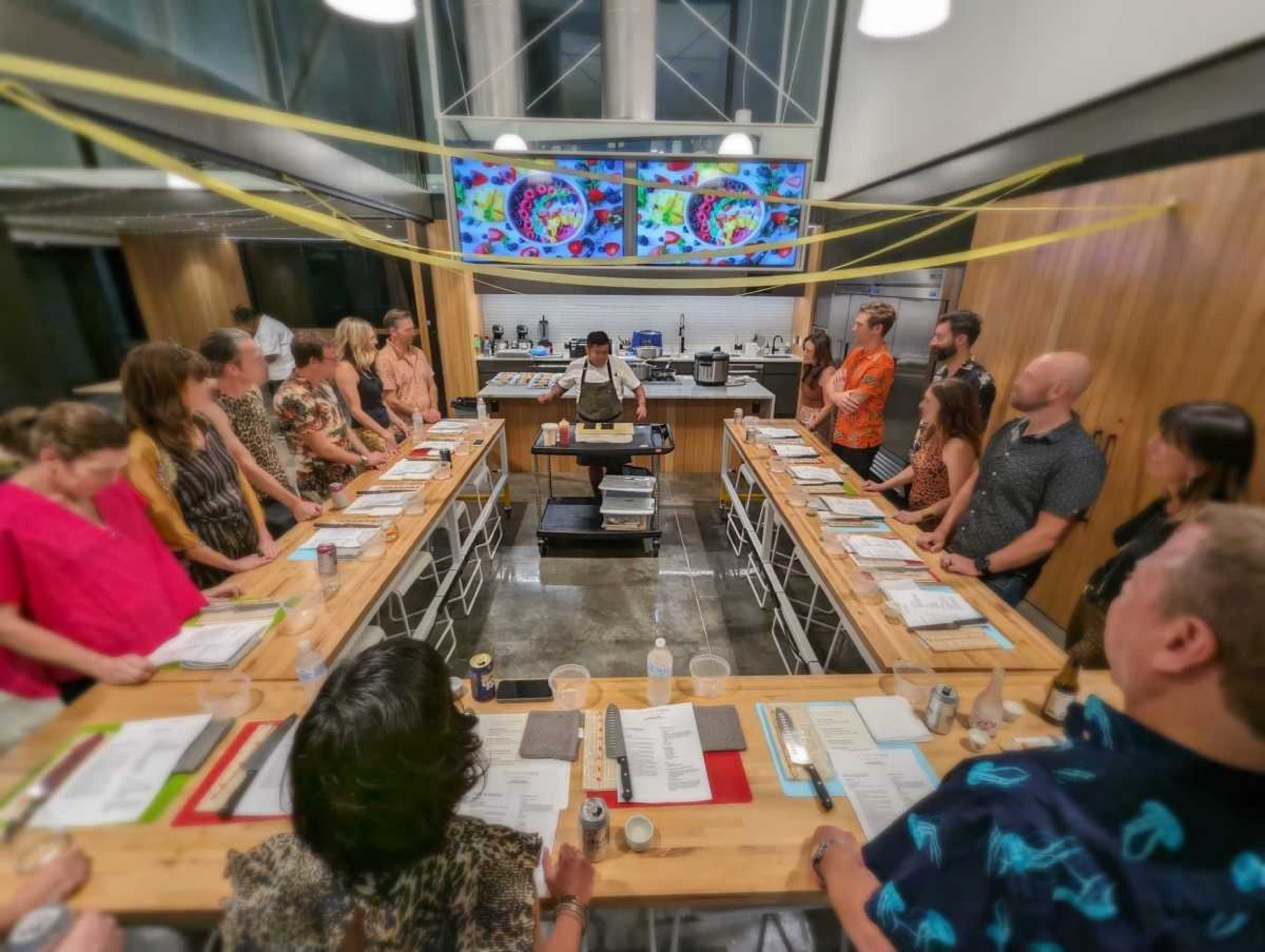 A group of people is gathered around a central cooking station in a modern kitchen, watching a chef prepare a meal.