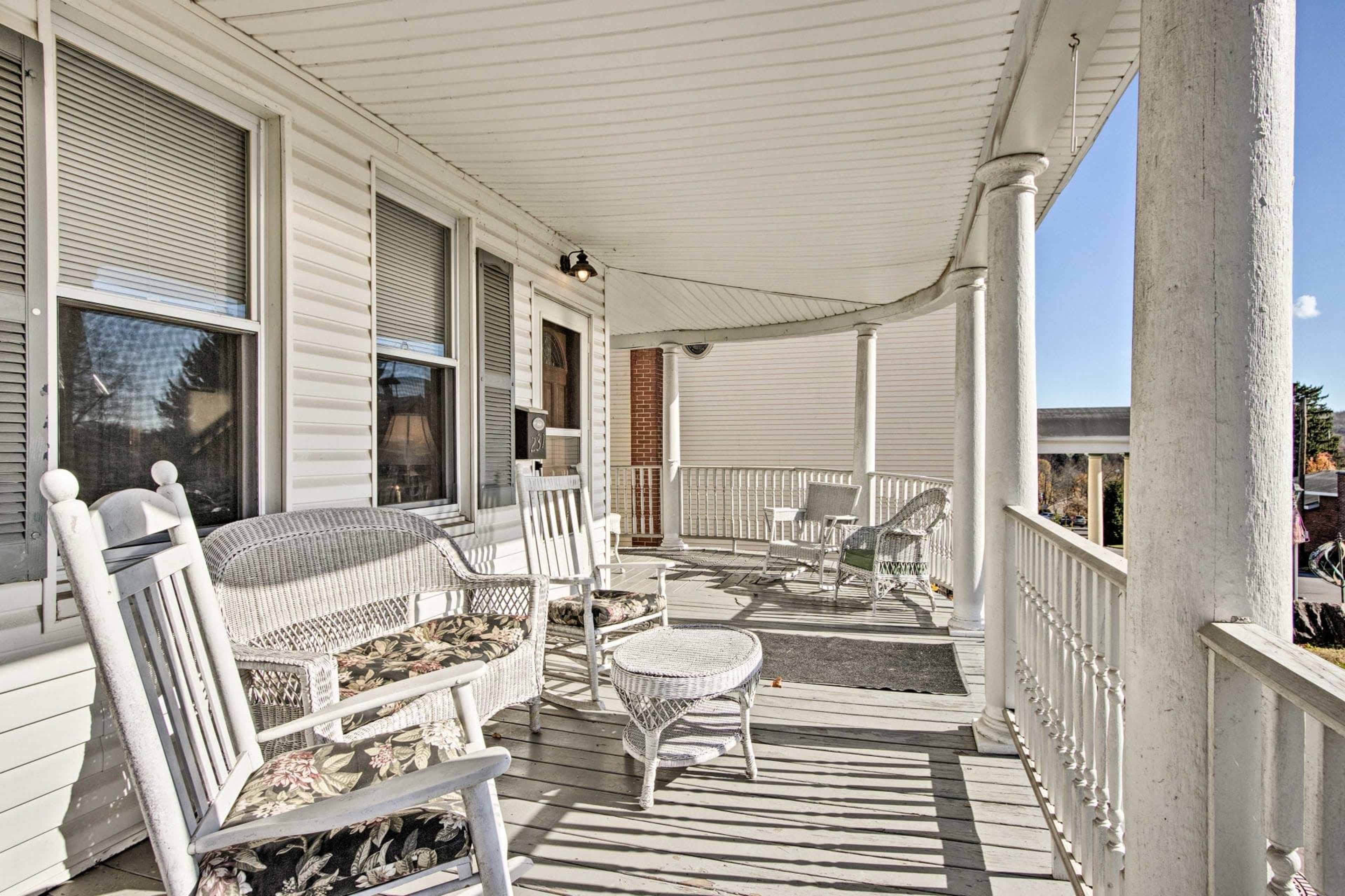 The image shows a covered porch with white wicker furniture, including rocking chairs and a small table, overlooking a clear sky.