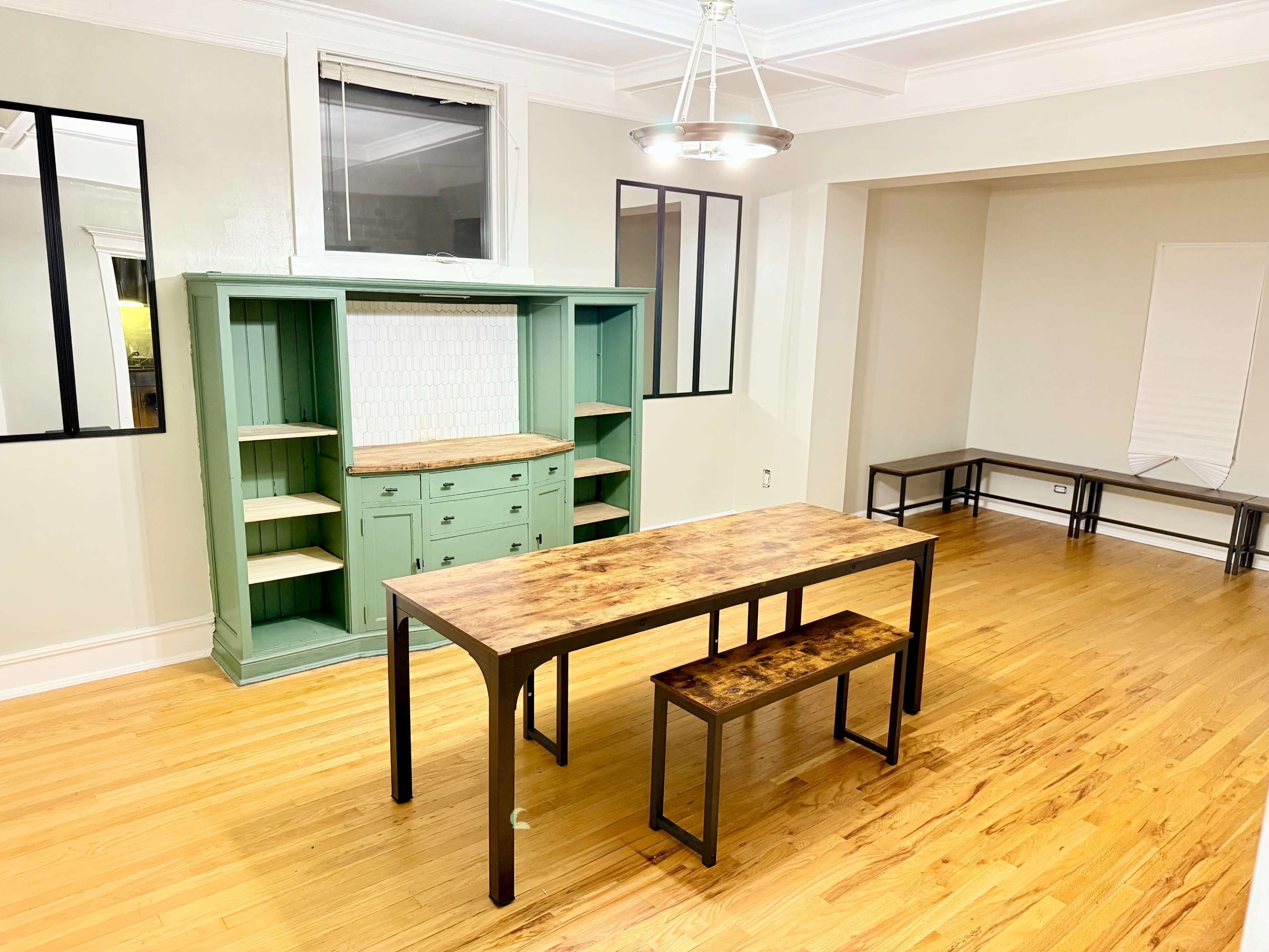 A wooden table with two stools is positioned in a well-lit room featuring a green cabinet and minimal furniture.