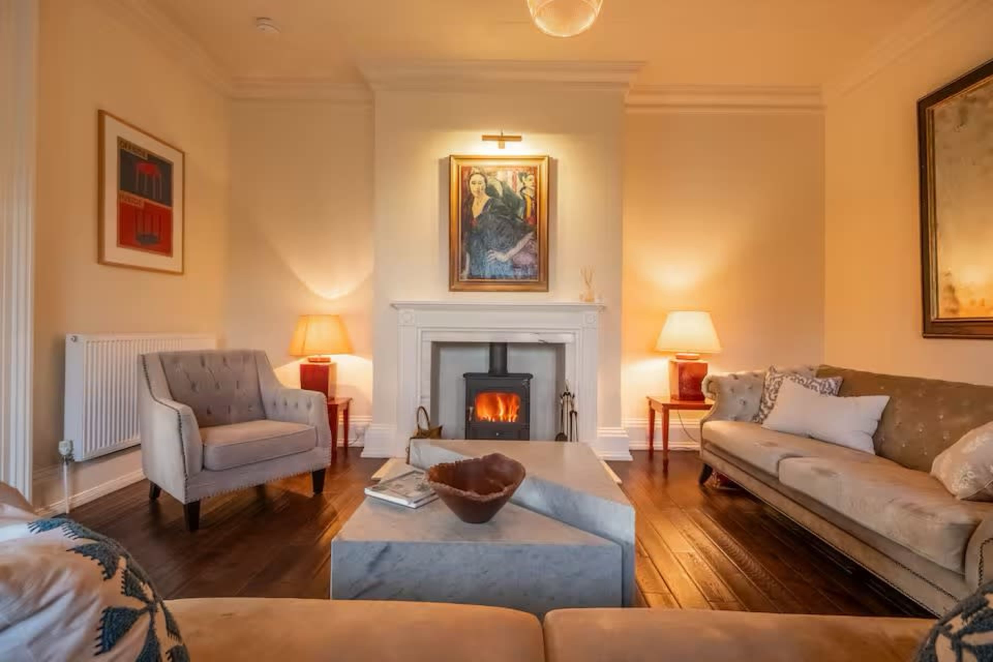 The living room features a fireplace with a painting above it, two side tables with lamps, and a central coffee table on a wooden floor.