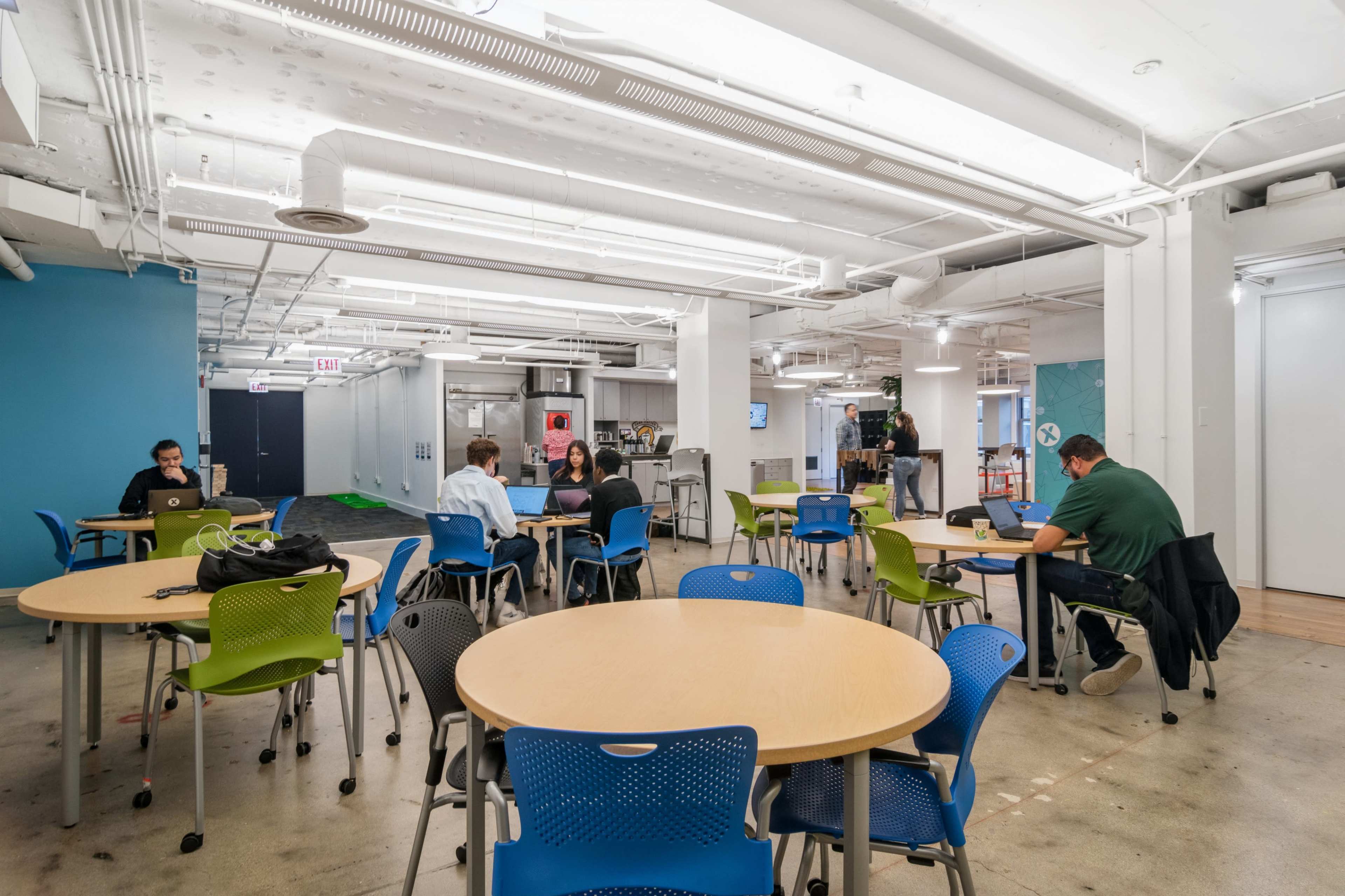 A spacious office area with several round tables, chairs in different colors, and individuals working at their laptops.