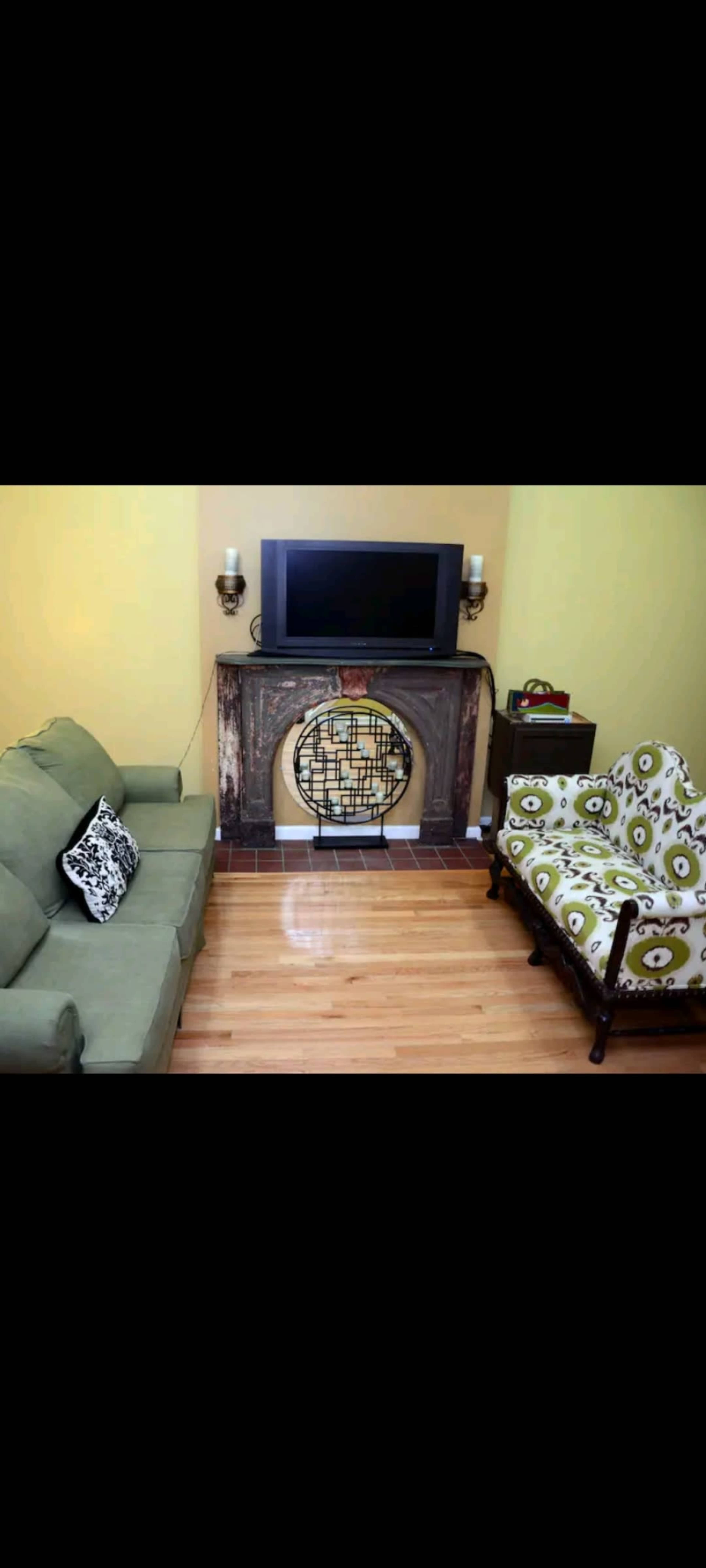 A living room with a green sofa, a patterned armchair, a fireplace with a round decorative screen, and a wall-mounted television.