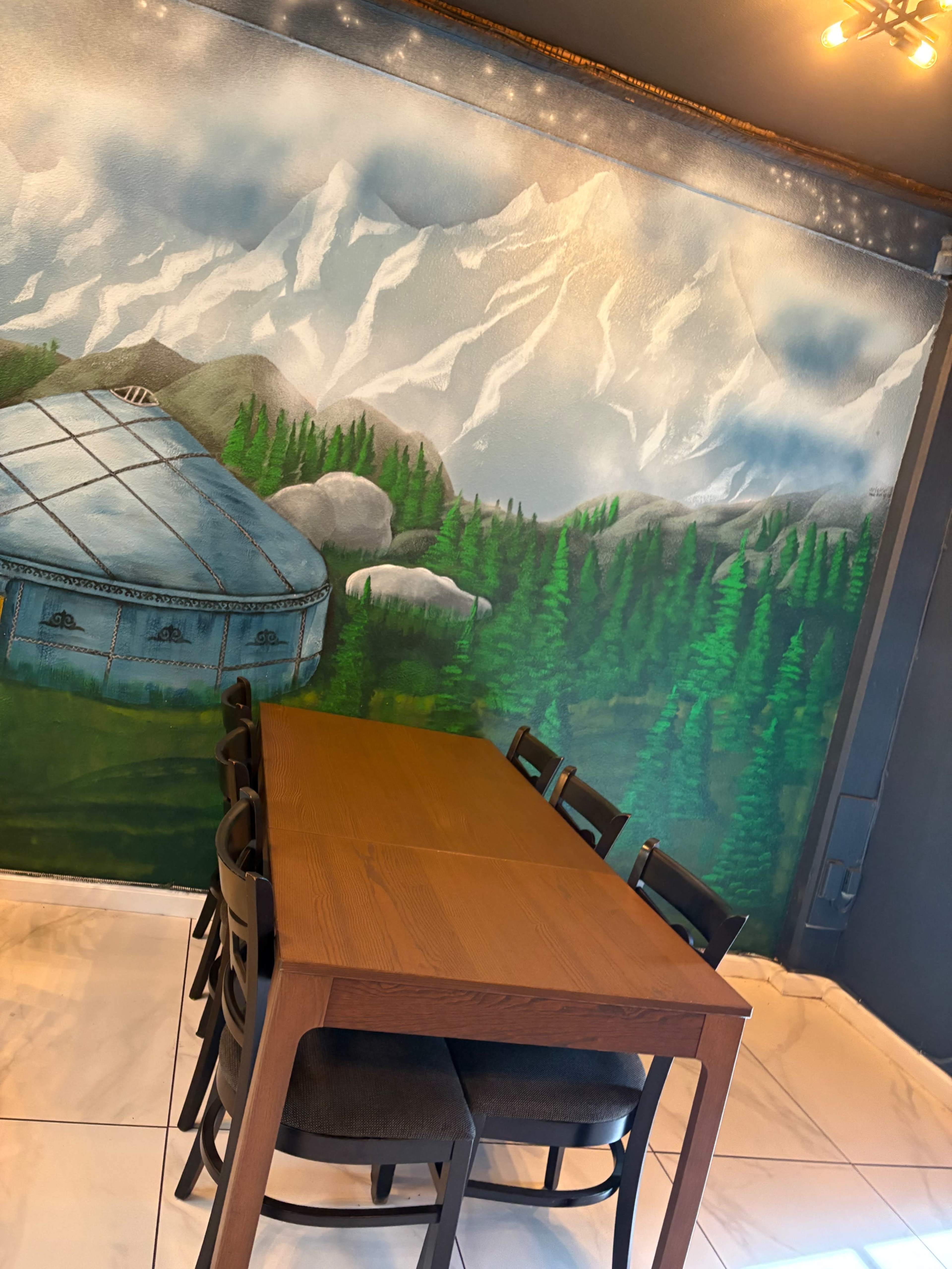 The image shows a wooden dining table surrounded by black chairs in front of a mural depicting mountains and trees.