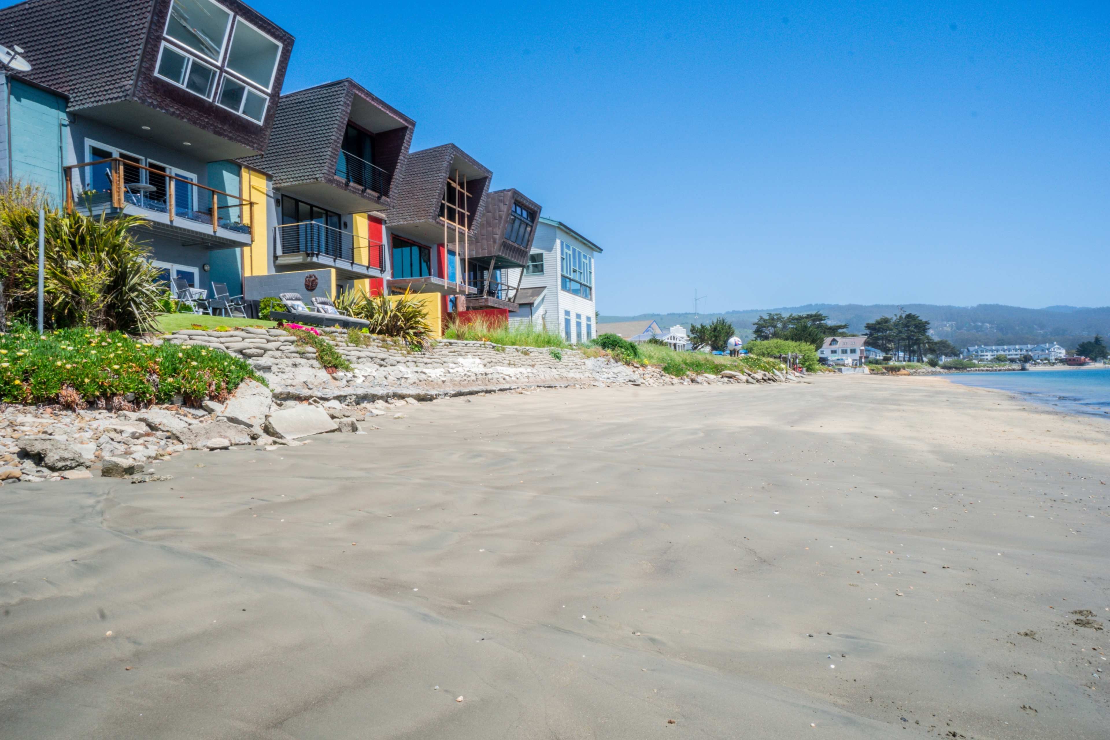 A line of colorful houses is situated along a sandy beach with calm waters and clear blue skies.