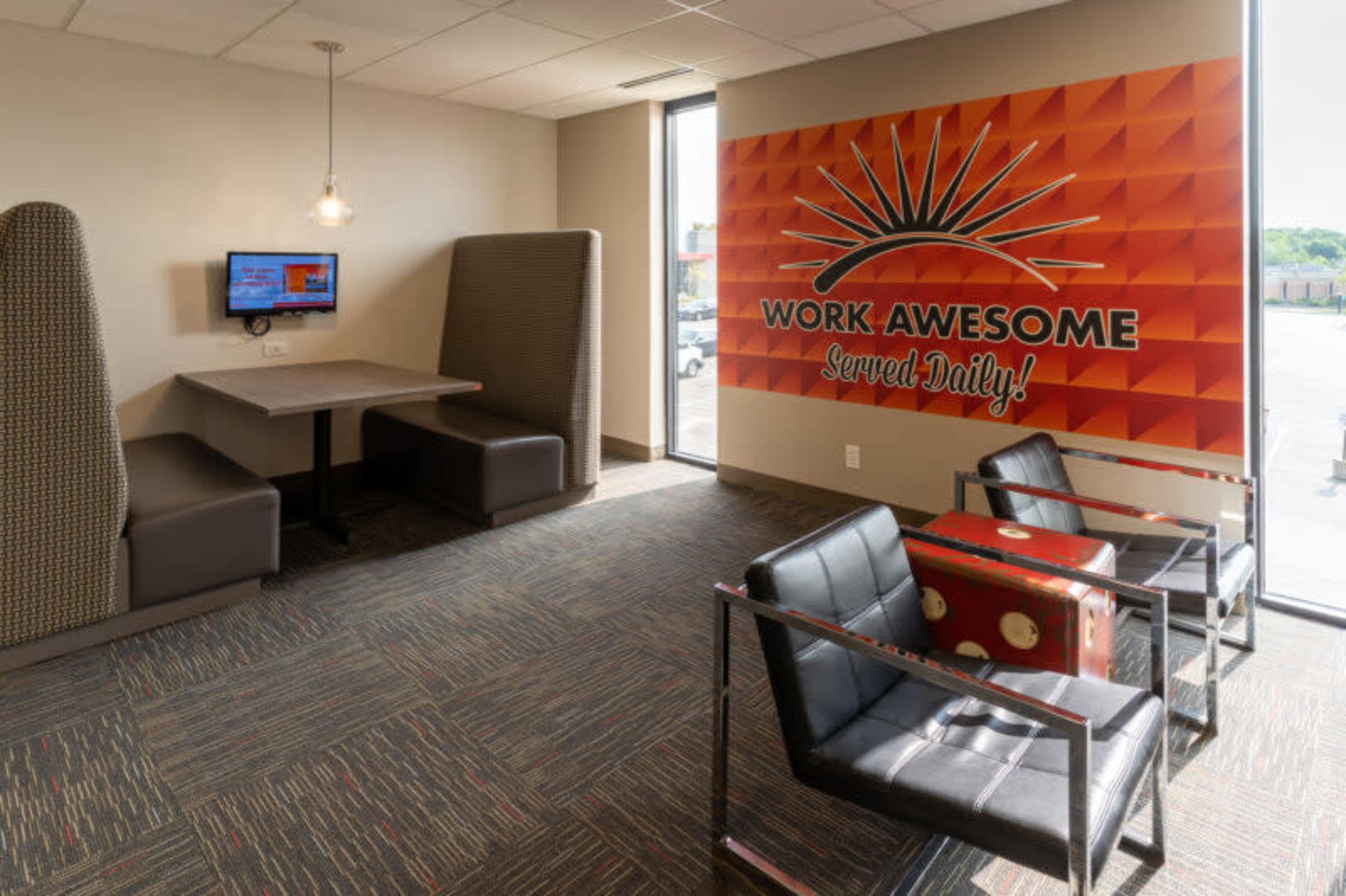 The image shows a modern workspace with seating arrangements, a large wall mural that reads "WORK AWESOME Served Daily!", and a small desk with a television set.