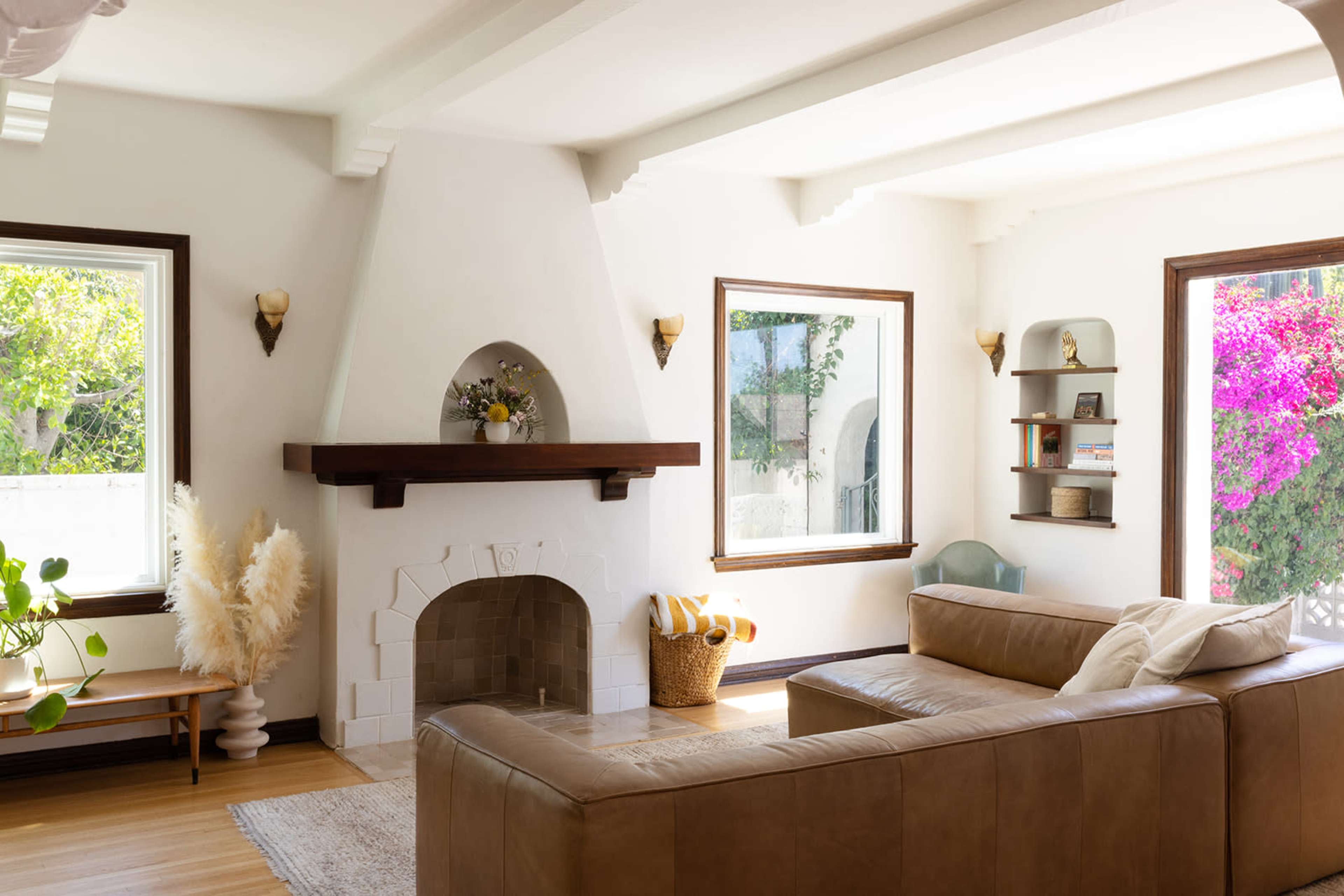 A spacious living room featuring a brown leather sectional sofa, an arched fireplace with a mantel, large windows, and shelves displaying decor, surrounded by greenery and pink flowers outside.