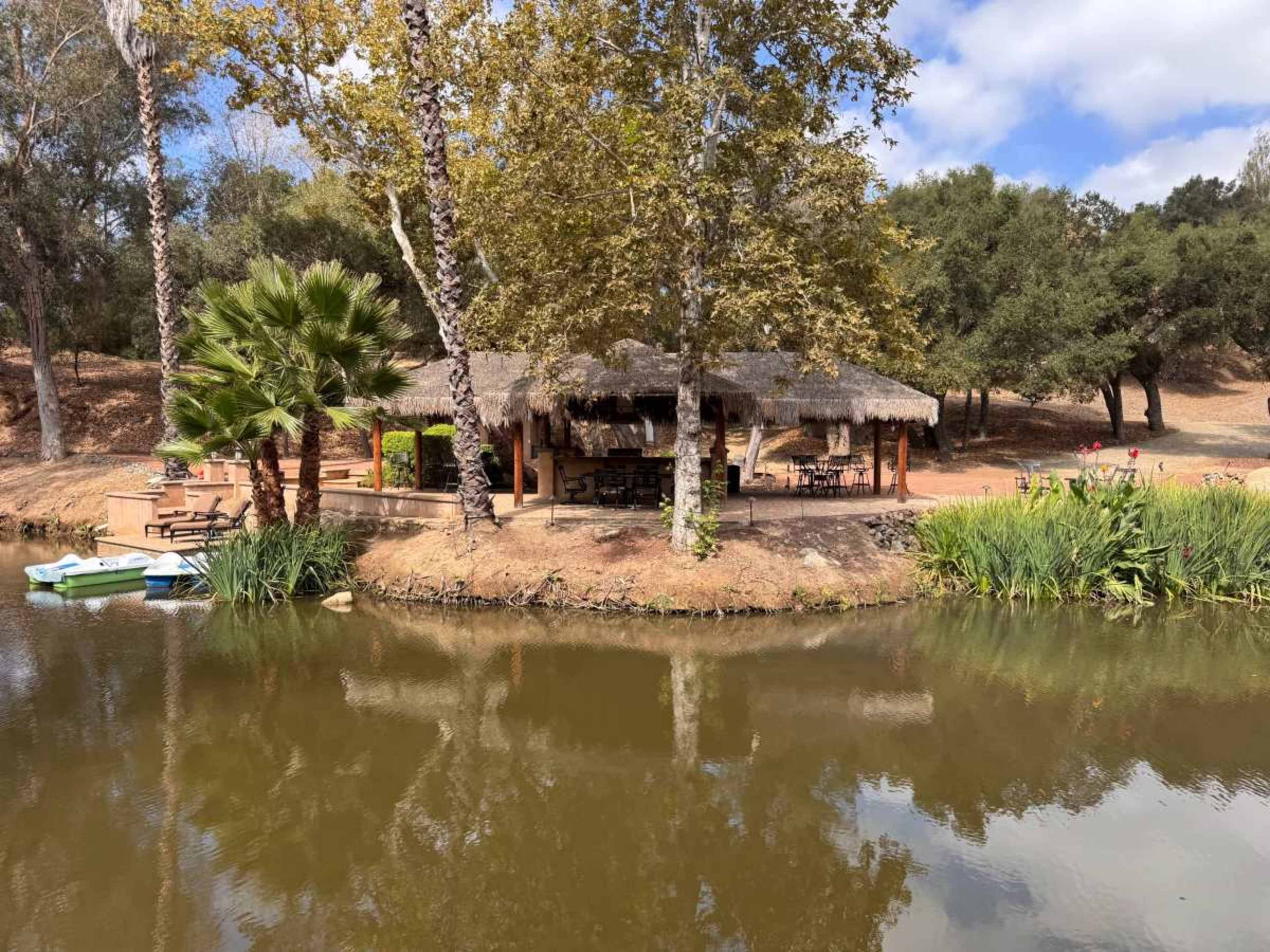 A thatched-roof structure sits along the edge of a serene pond surrounded by trees and greenery.