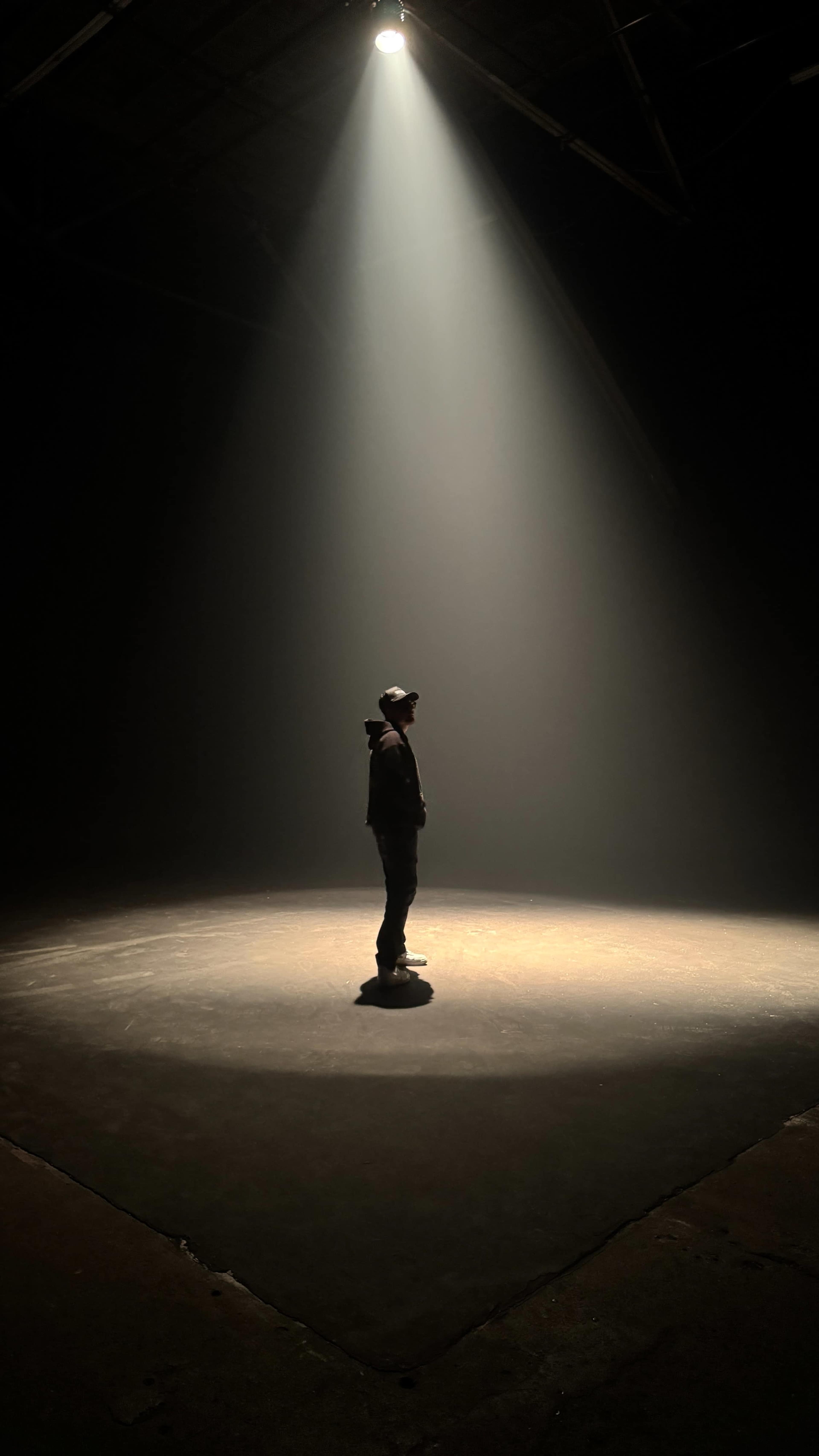 A person stands in a spotlight on a dark stage, surrounded by shadows.