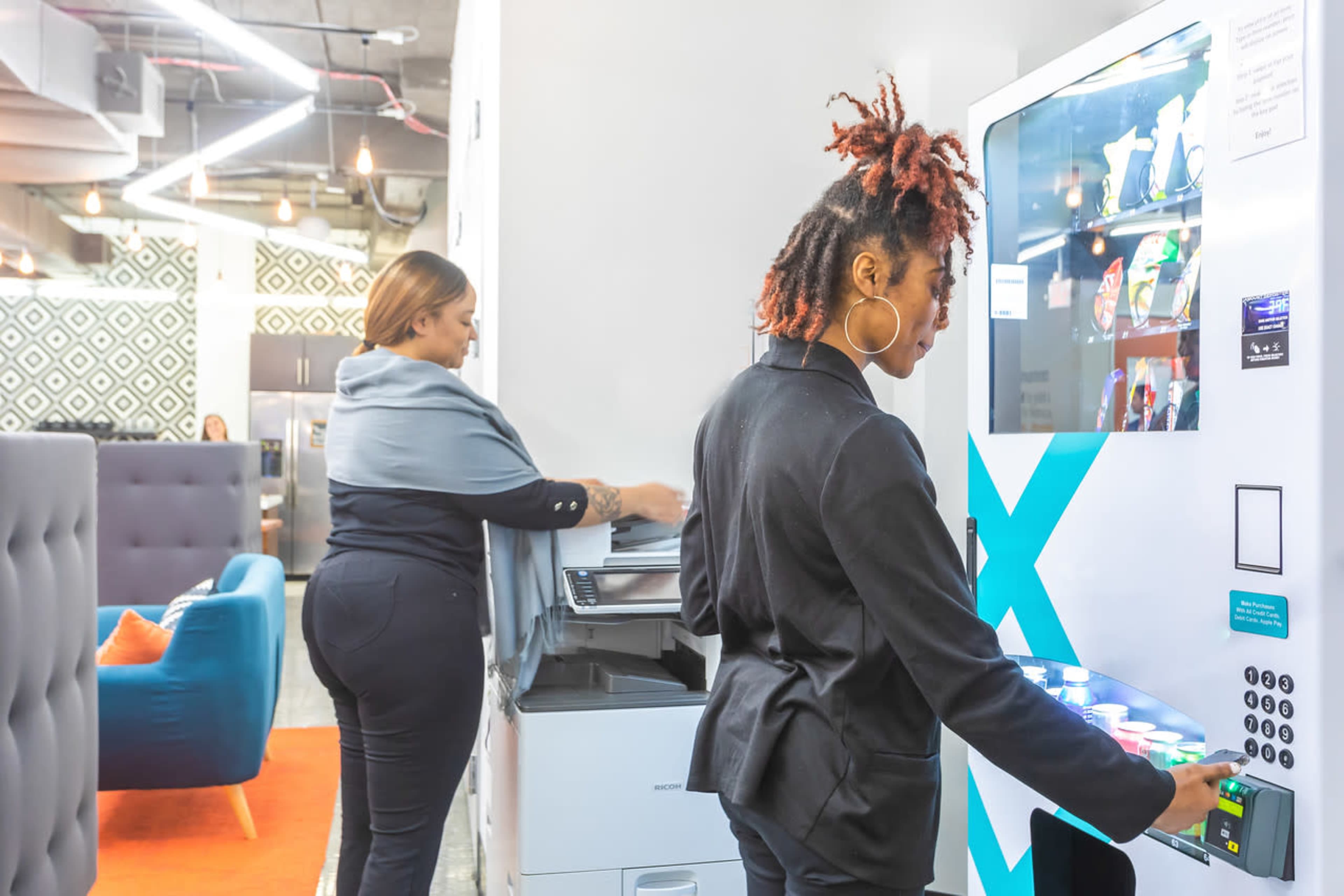Two women are in an office space, with one woman using a vending machine while the other sorts papers near a printer.