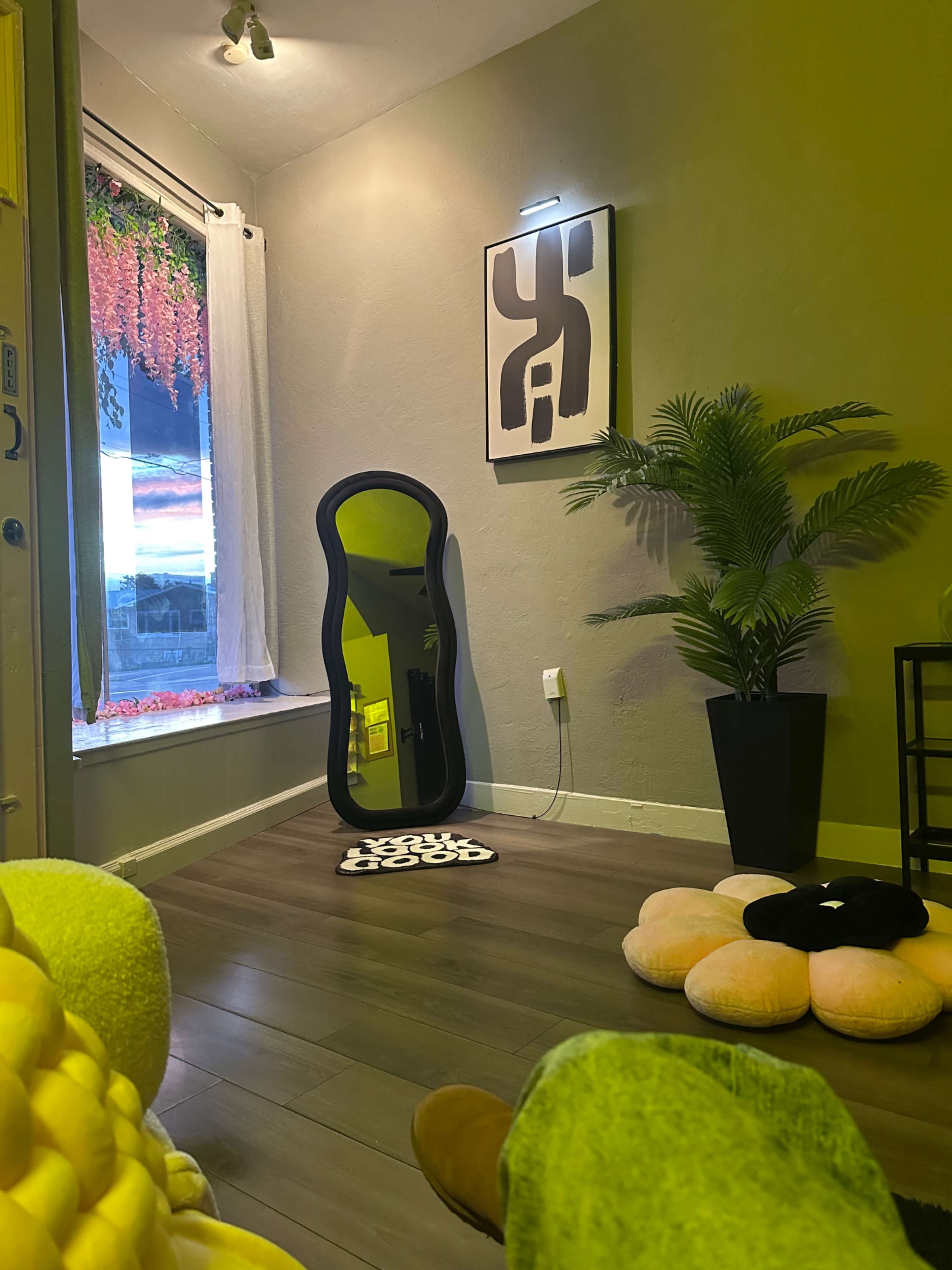 The image features a cozy room with a large mirror, a potted plant, and colorful floor cushions, illuminated by natural light from a window.