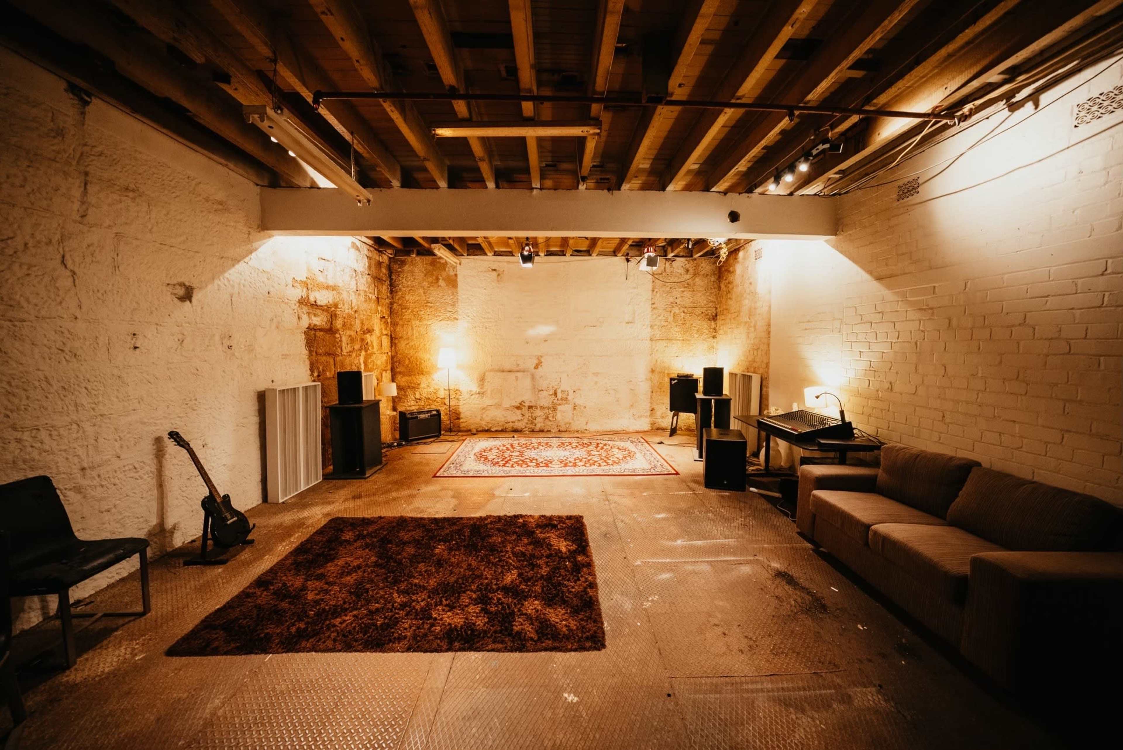 A spacious, empty basement room features a patterned rug, a sofa, a guitar, and speakers arranged along the walls.