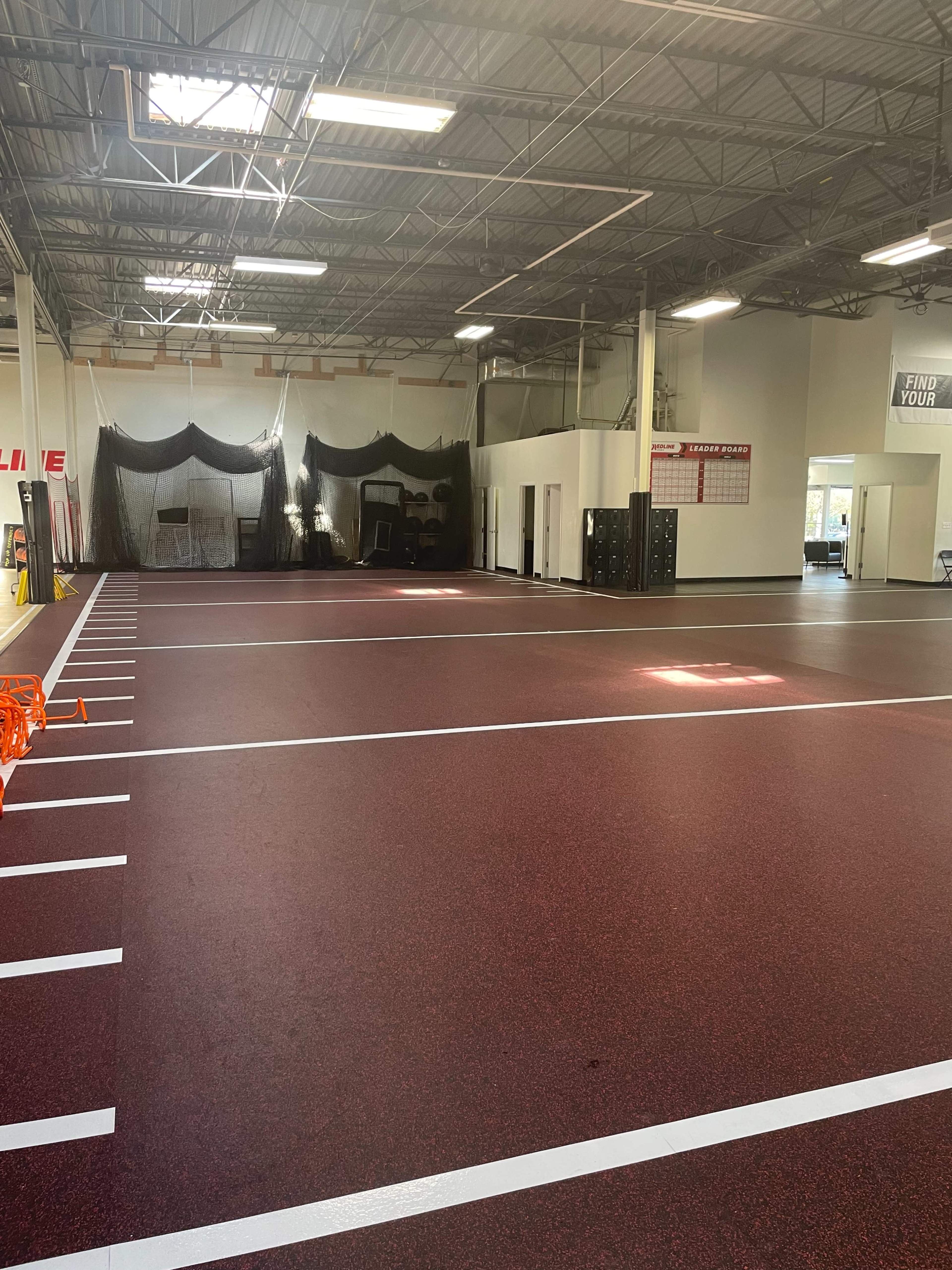 The image shows a spacious indoor training facility with a rubberized red floor and marked lanes, illuminated by overhead lights.