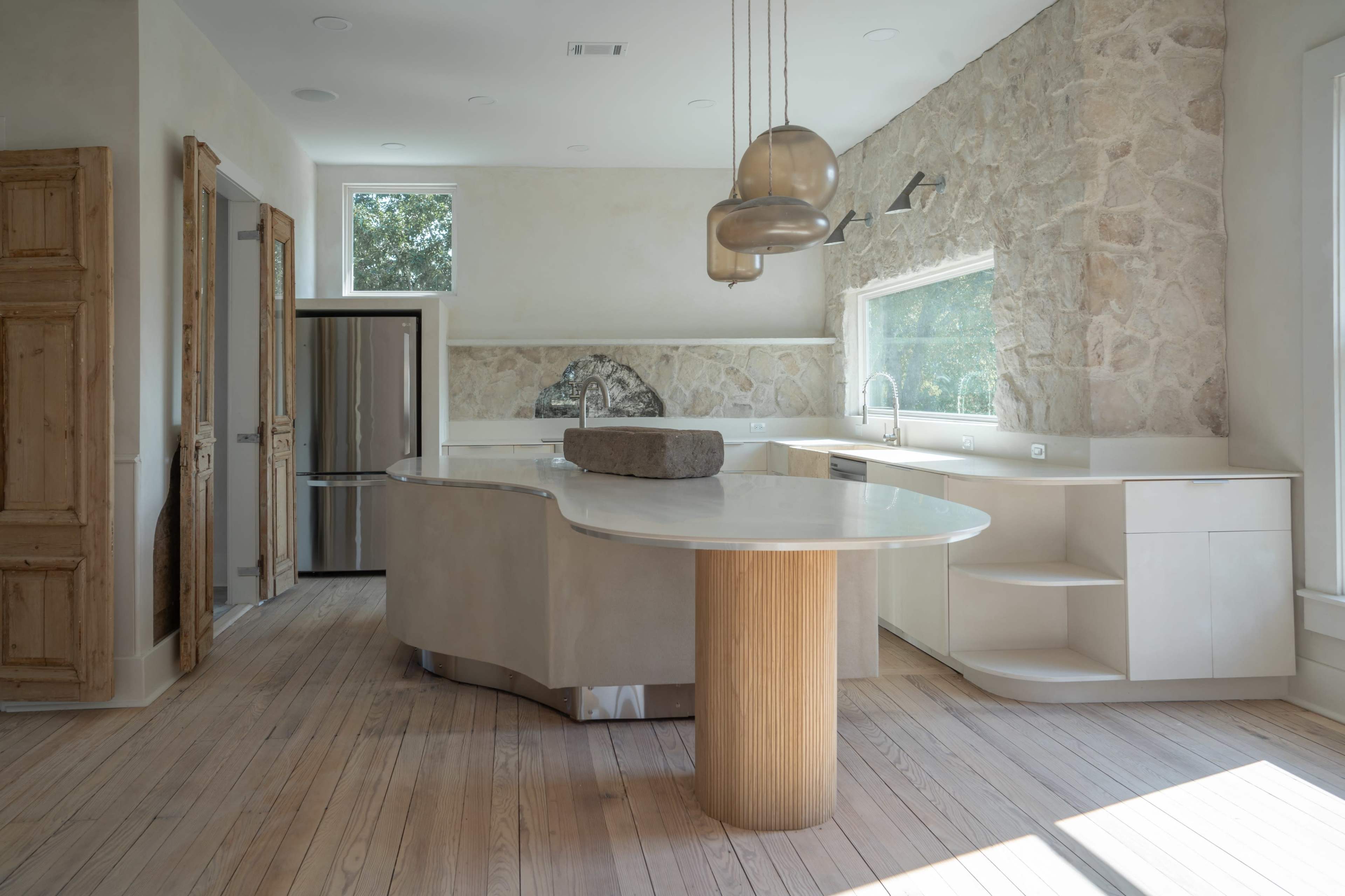 A modern kitchen with a large, curved marble island, wooden flooring, and stone walls, featuring sleek cabinetry and large windows.
