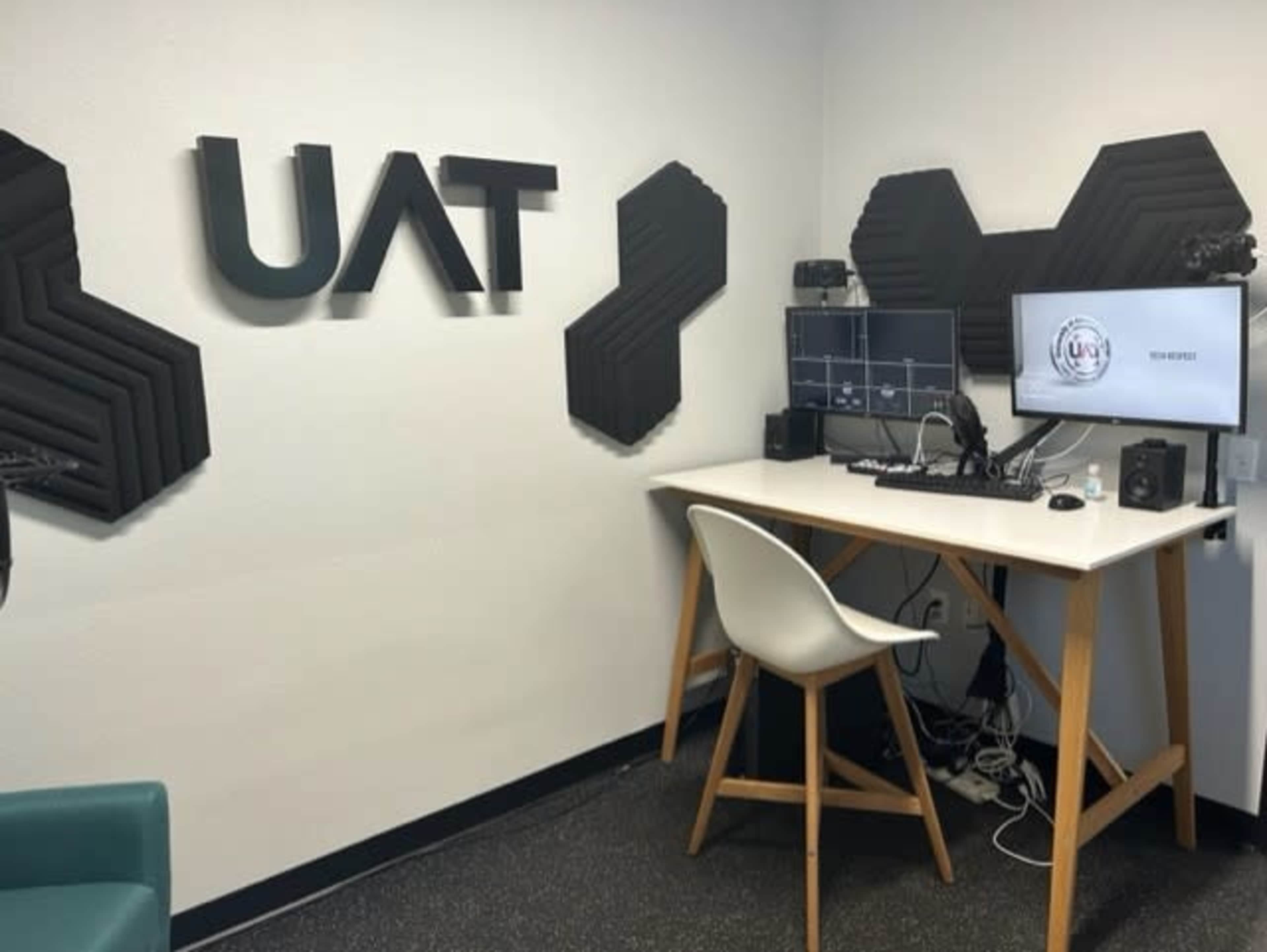 A modern workspace featuring a desk with dual monitors, a webcam, and audio equipment against a wall with acoustic panels and the letters "UAT" displayed prominently.