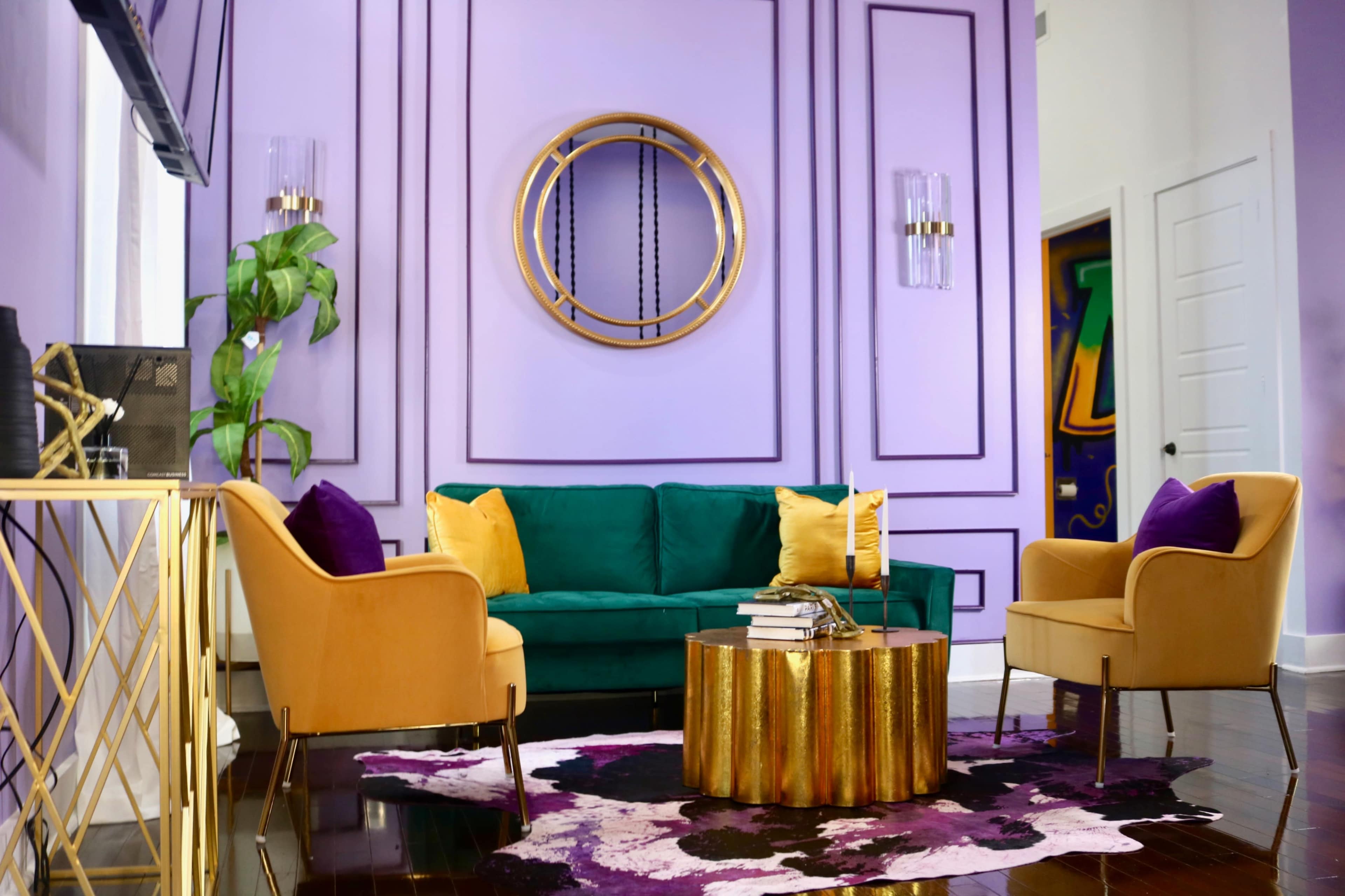 A vibrant living room features a green sofa and two light-colored chairs around a gold and wood coffee table, with purple and yellow accents against a lavender wall.