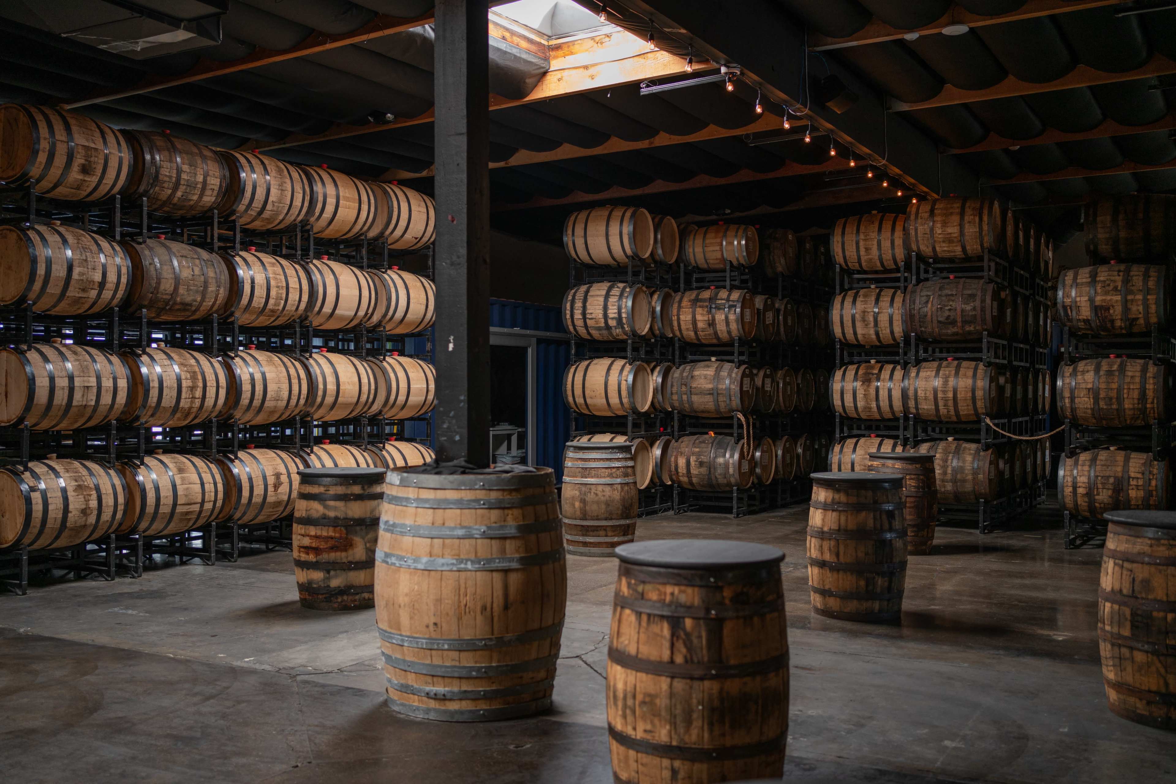 The image shows a room filled with stacked wooden barrels on metal racks, with a few standalone barrels placed on the floor.