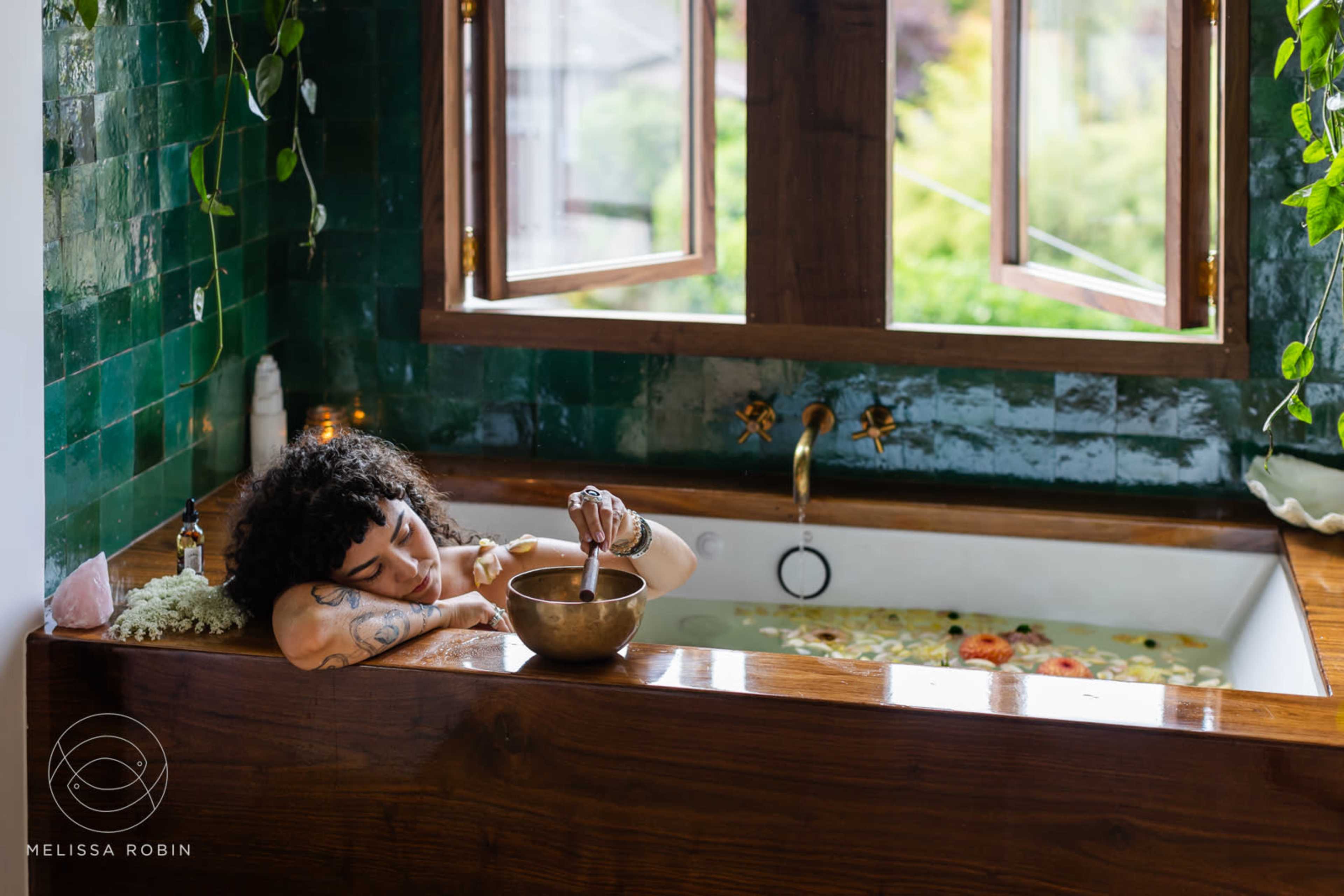 A person rests beside a bathtub filled with flowers, while holding a bowl in a room with large windows and green tiles.