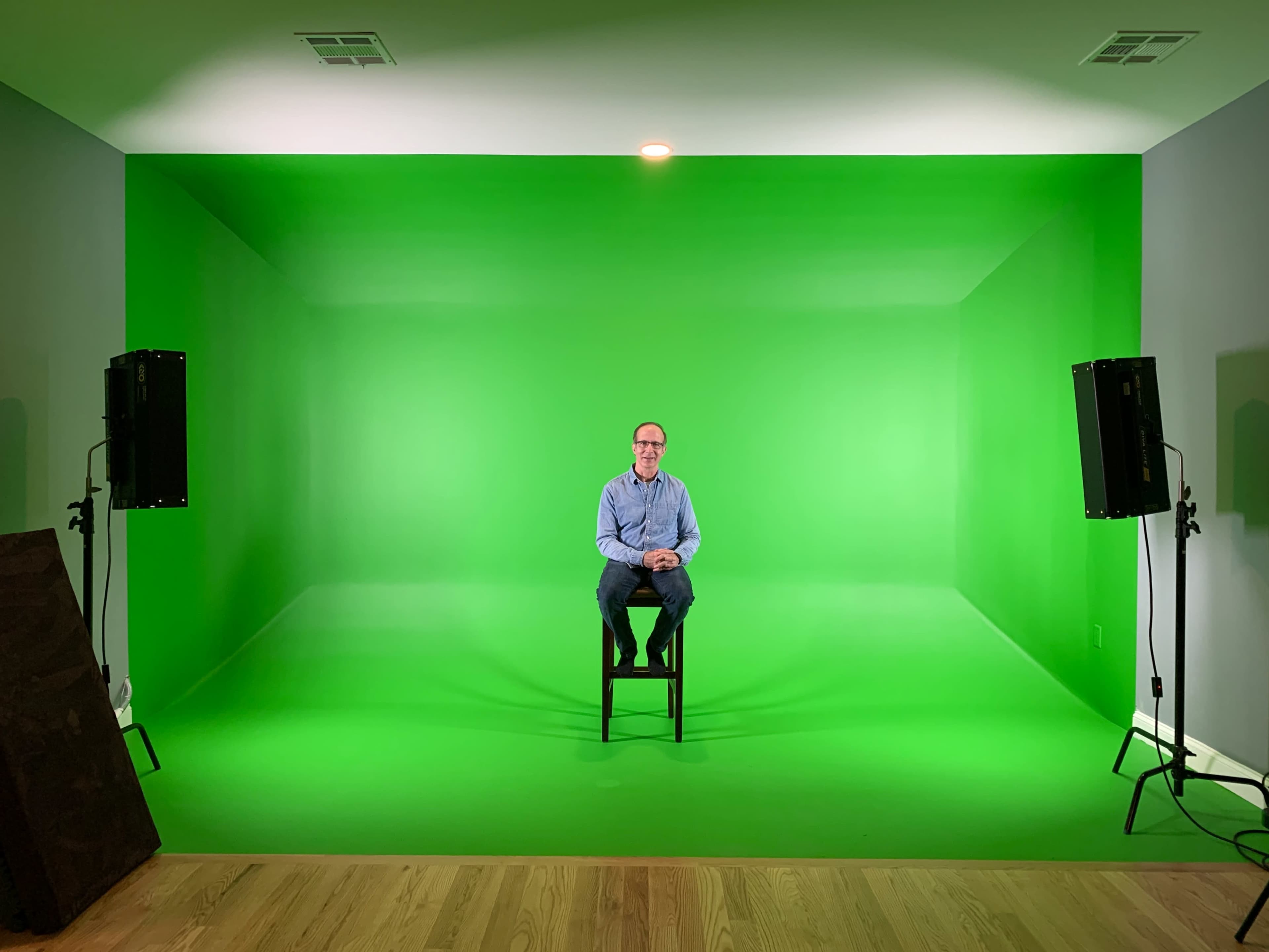 A man sits on a stool in front of a green screen, surrounded by two lights, in a spacious room with wooden flooring.