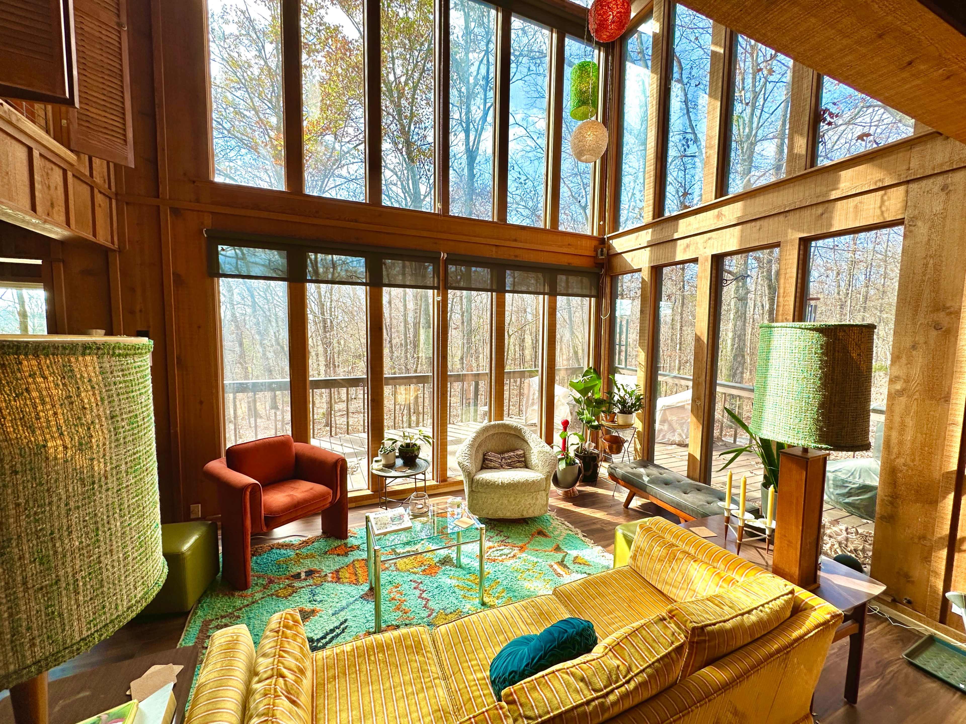 A spacious wooden living room with large windows overlooking a forest, featuring modern furniture and decorative plants.