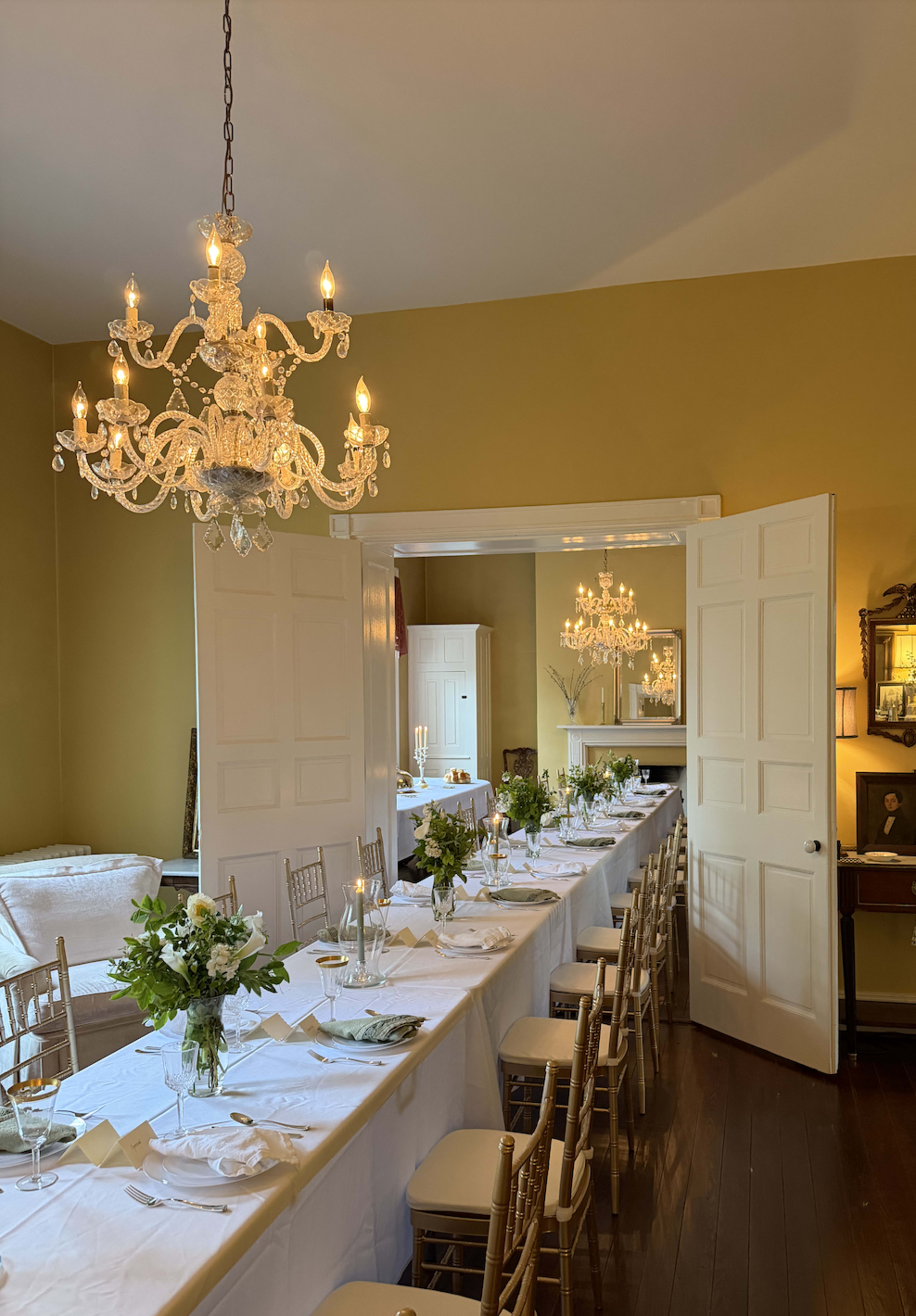 A long dining table is set with white linens and flowers, illuminated by a chandelier in a warmly colored room.