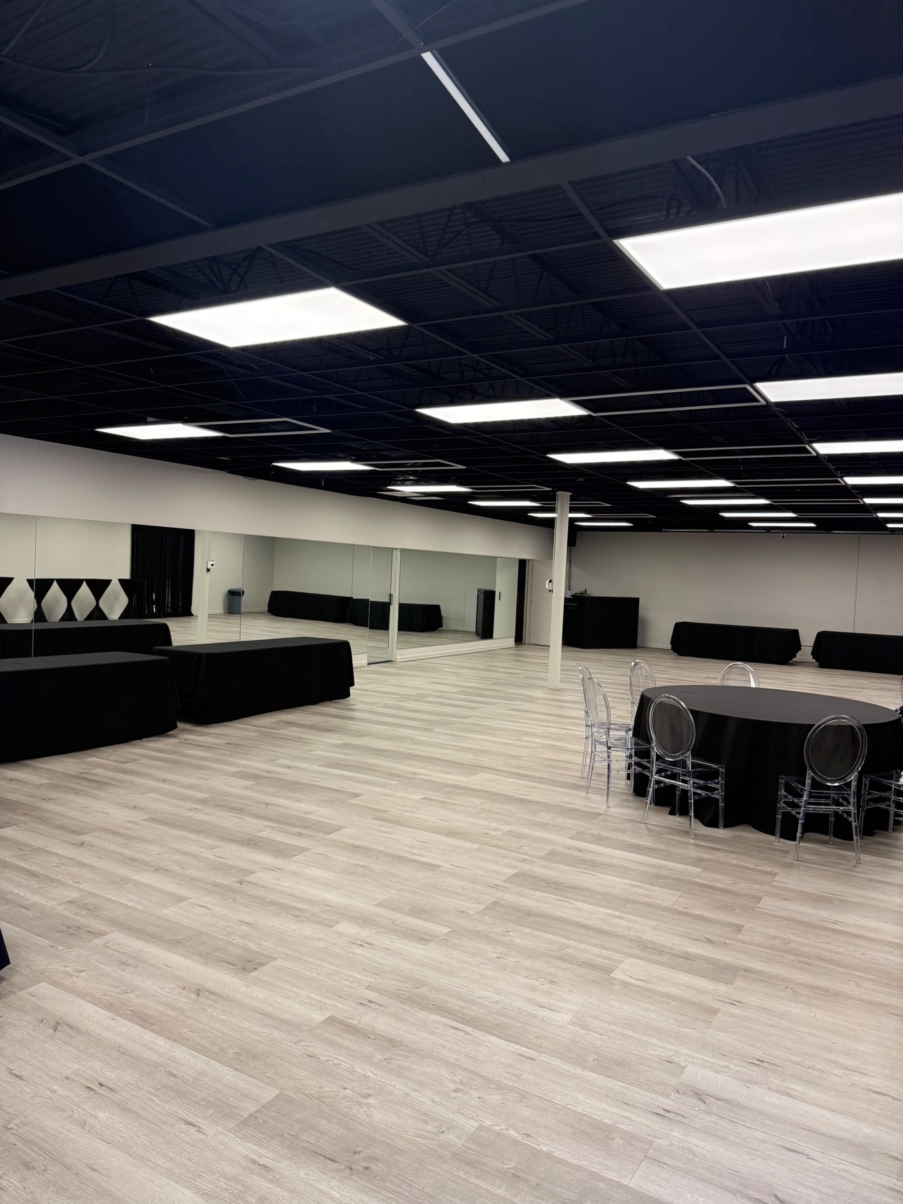 The image shows a spacious, empty event room with light wood flooring, black tables along the walls, and a round table with transparent chairs in the foreground.