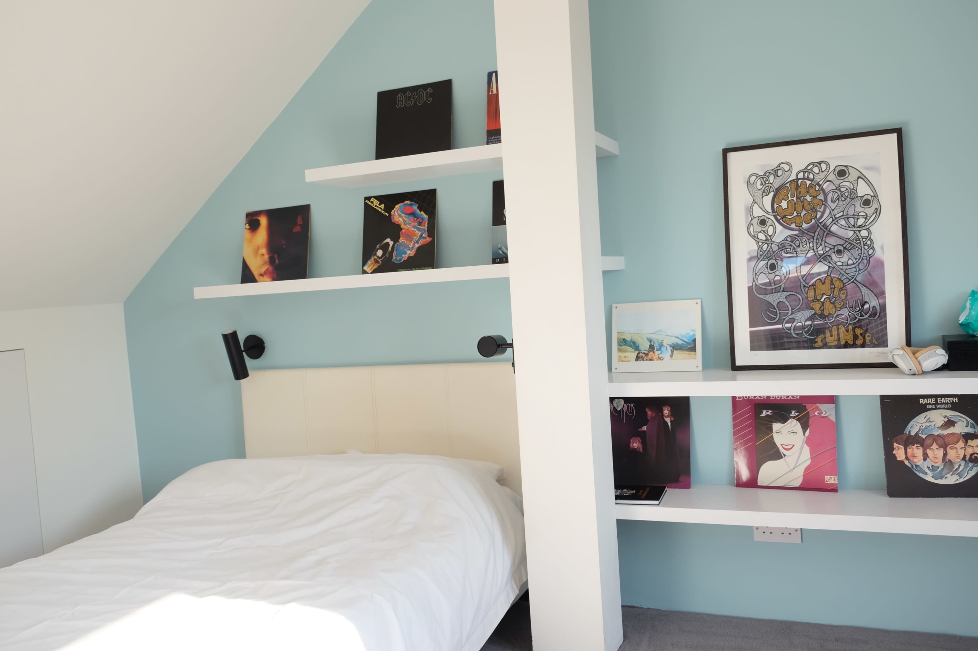 A bedroom with a white bed against a light blue wall, featuring floating shelves displaying framed art and vinyl records.