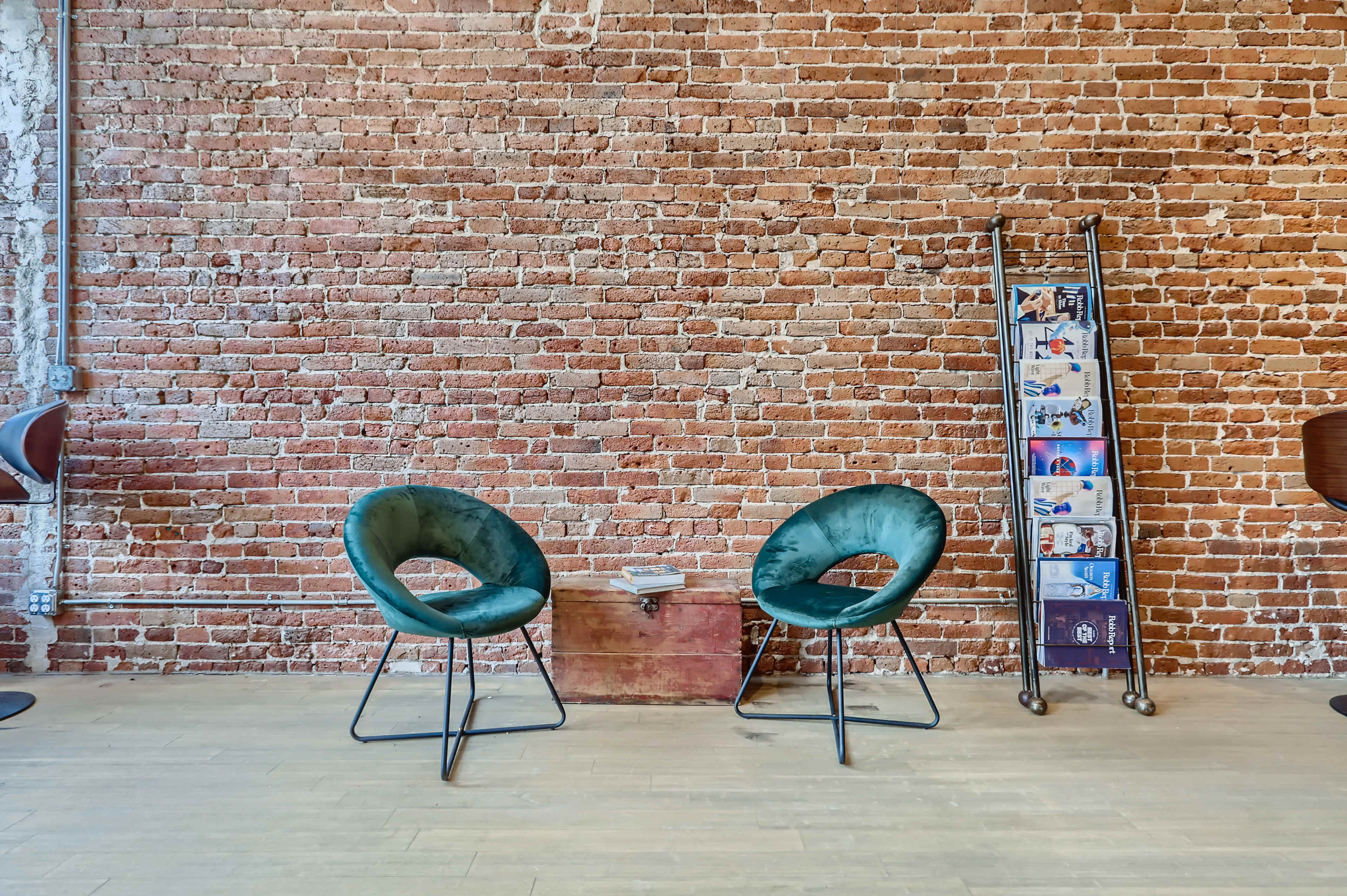 Two green chairs facing each other in front of a brick wall, with a metal rack holding brochures nearby.