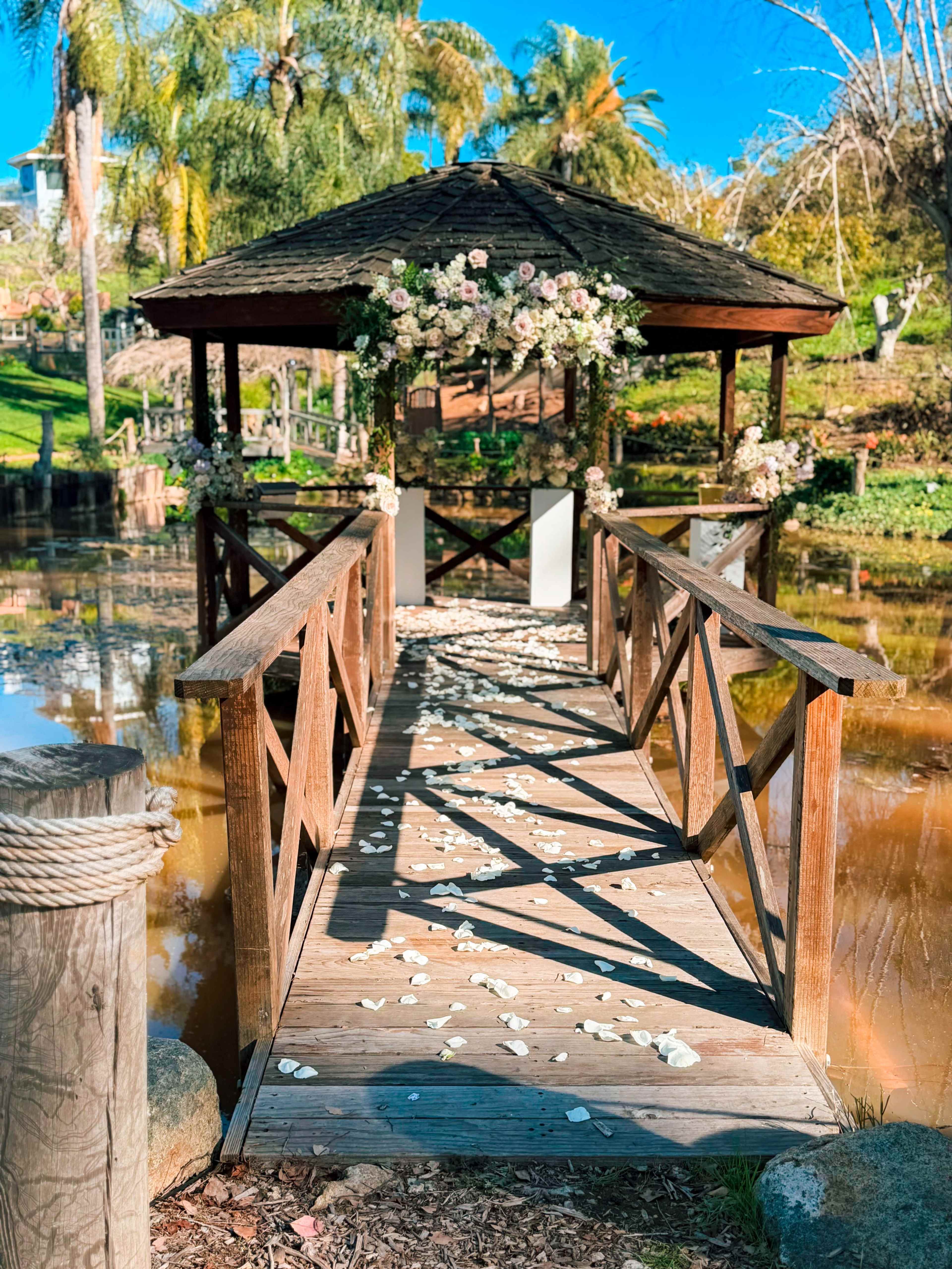 A wooden bridge leads to a gazebo adorned with flowers, with rose petals scattered across the walkway and a serene pond reflecting the surroundings.