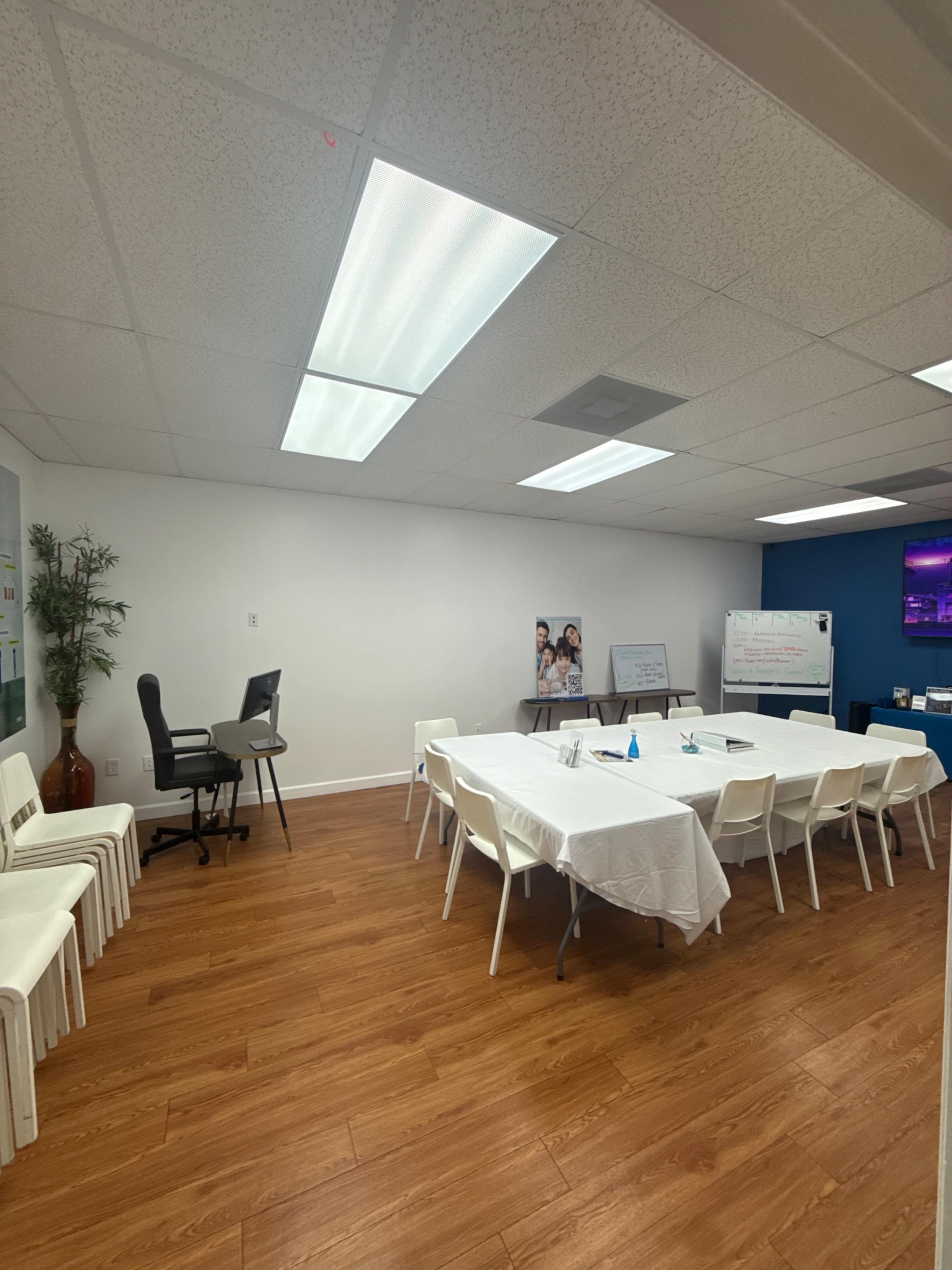 The image shows a conference room with a long table covered in a white tablecloth, surrounded by white chairs, and featuring a presentation area with a monitor and a black office chair.
