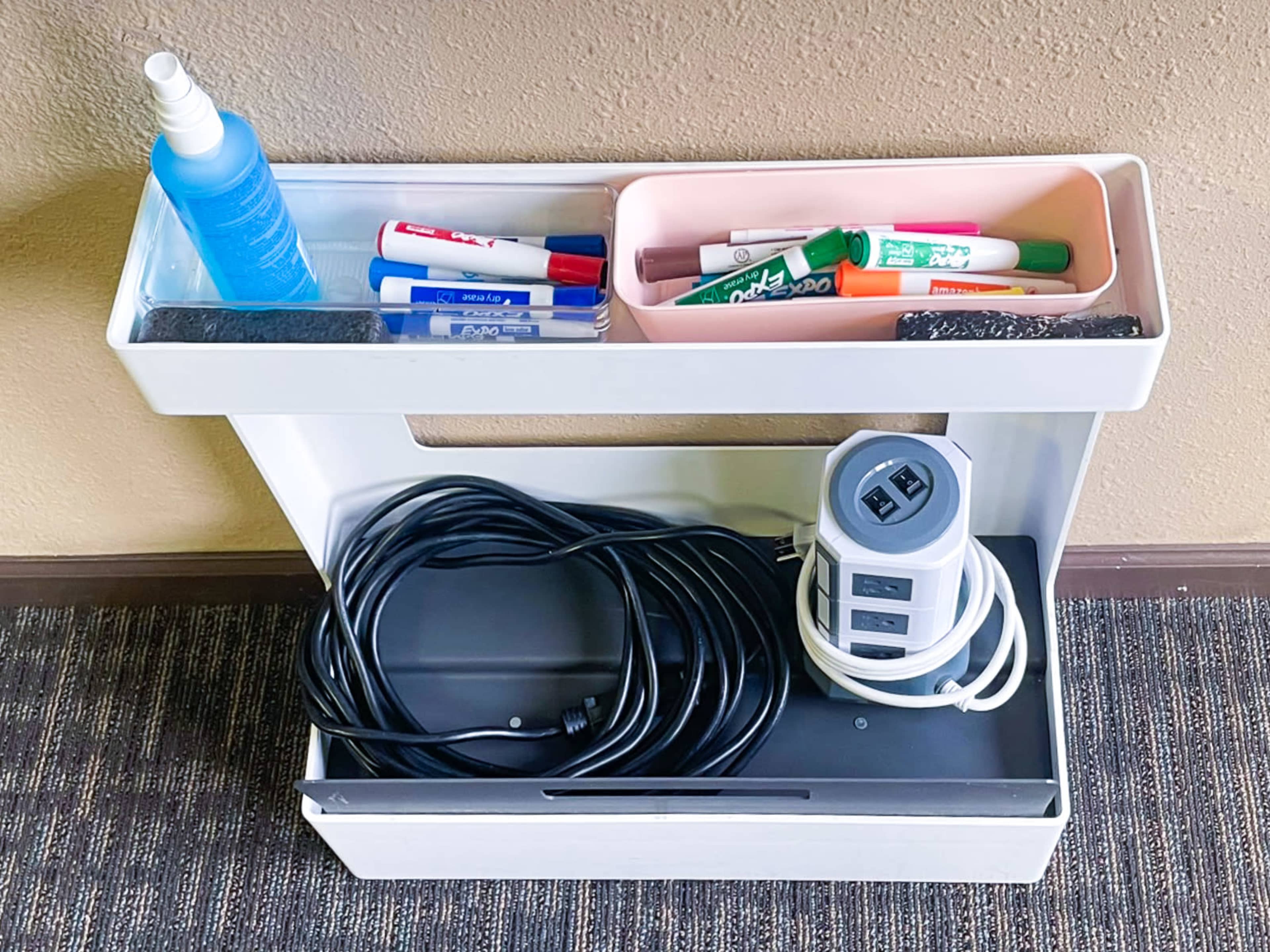A white storage cart with compartments holding cleaning spray, markers, and a power strip with multiple outlets, situated against a beige wall.