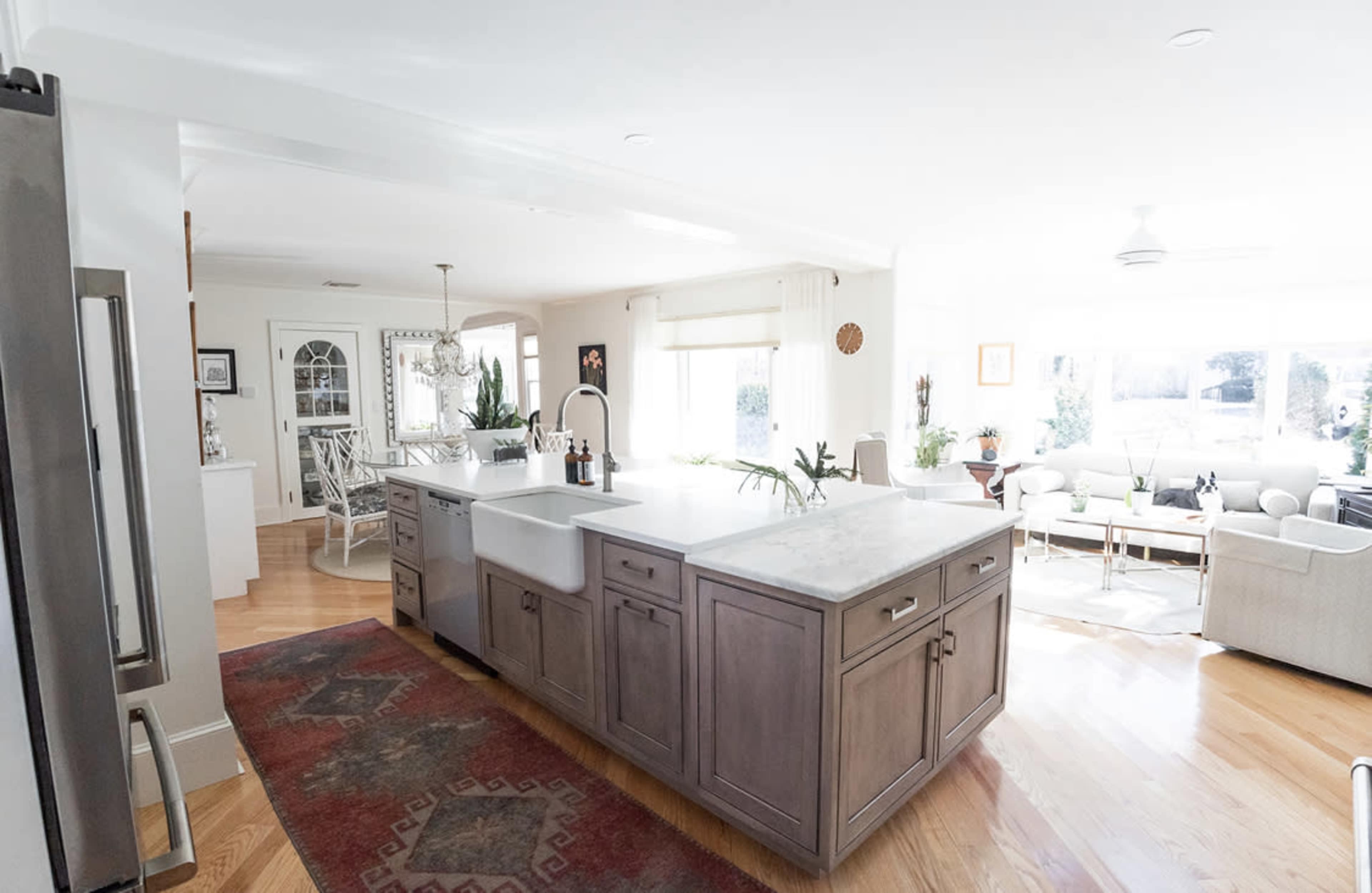 The image shows a bright, open kitchen with a large island, featuring a farmhouse sink and modern appliances, adjacent to a living area with windows.