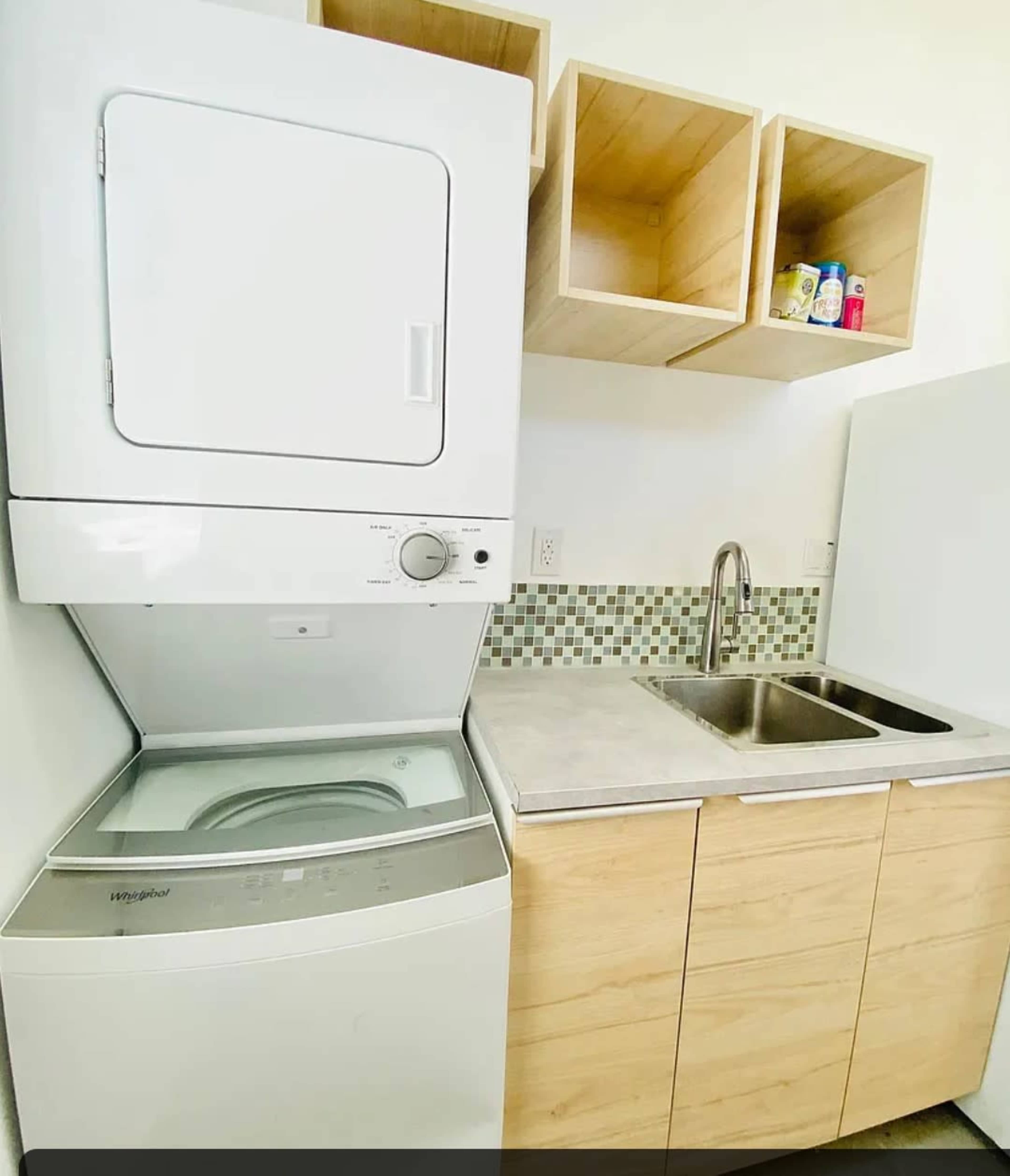 A stacked washer and dryer are positioned next to a sink with a countertop and wooden cabinets above it, featuring decorative storage boxes.