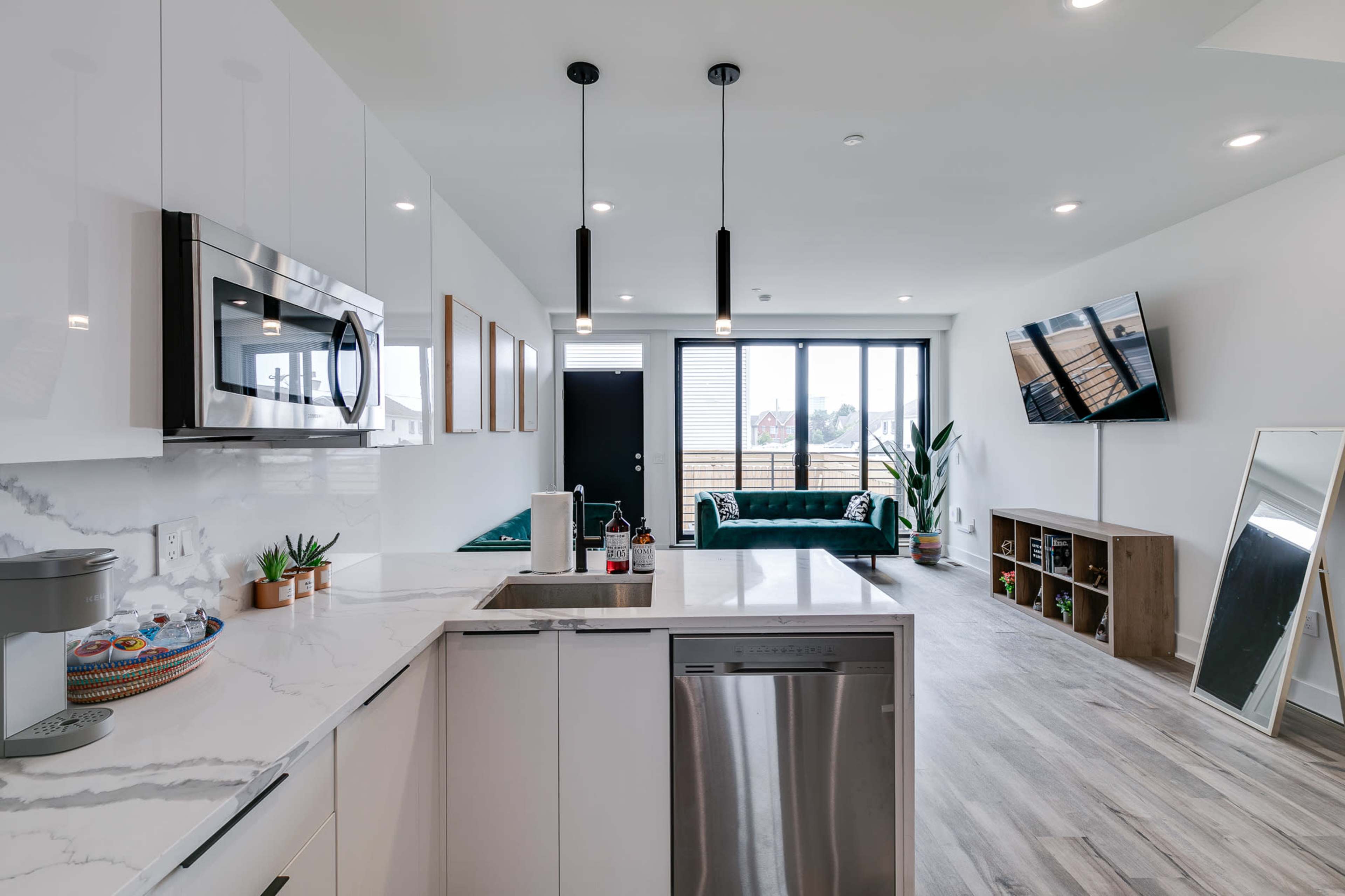 A modern kitchen area features a marble countertop, stainless steel appliances, and an open living space with large windows and plants.