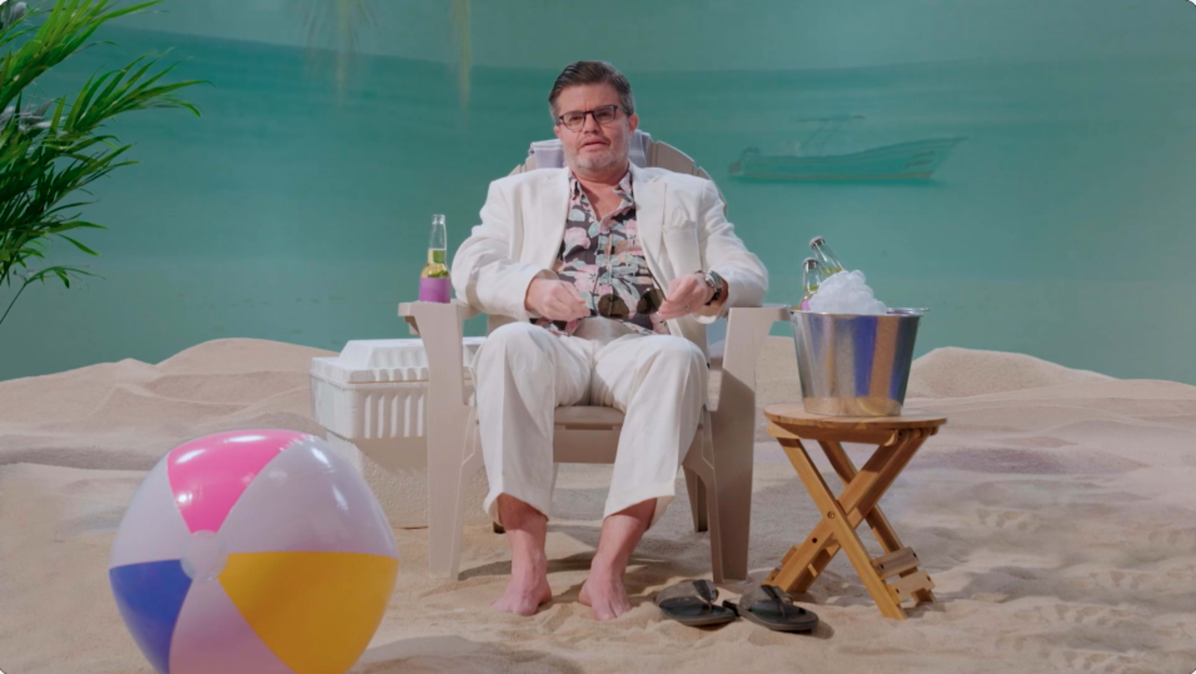 A man in a white suit sits in a beach chair on sandy ground, surrounded by a beach ball and a cooler, with a bucket of drinks nearby.