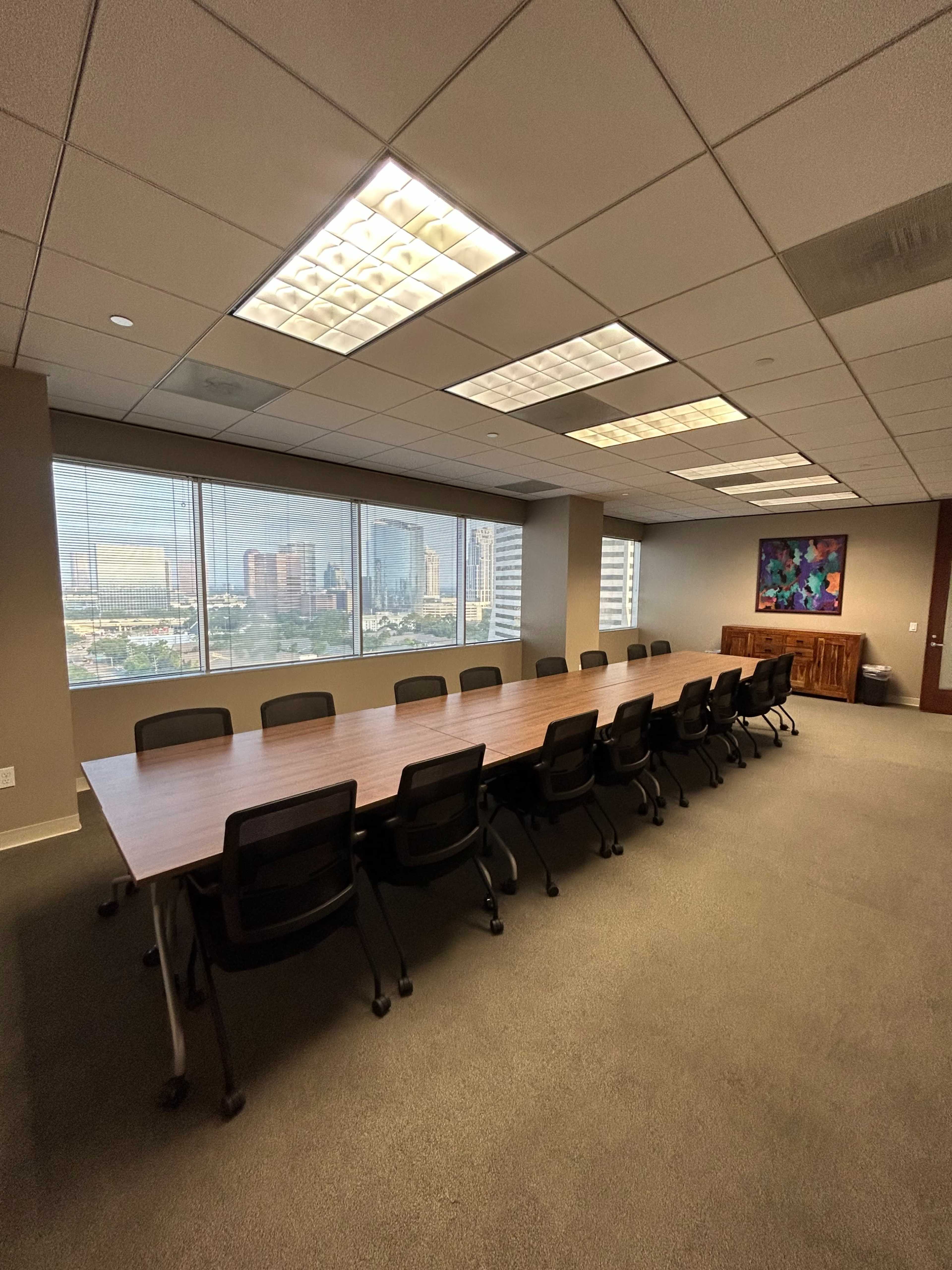 A large conference room features a long wooden table surrounded by black chairs, with large windows displaying a city skyline.