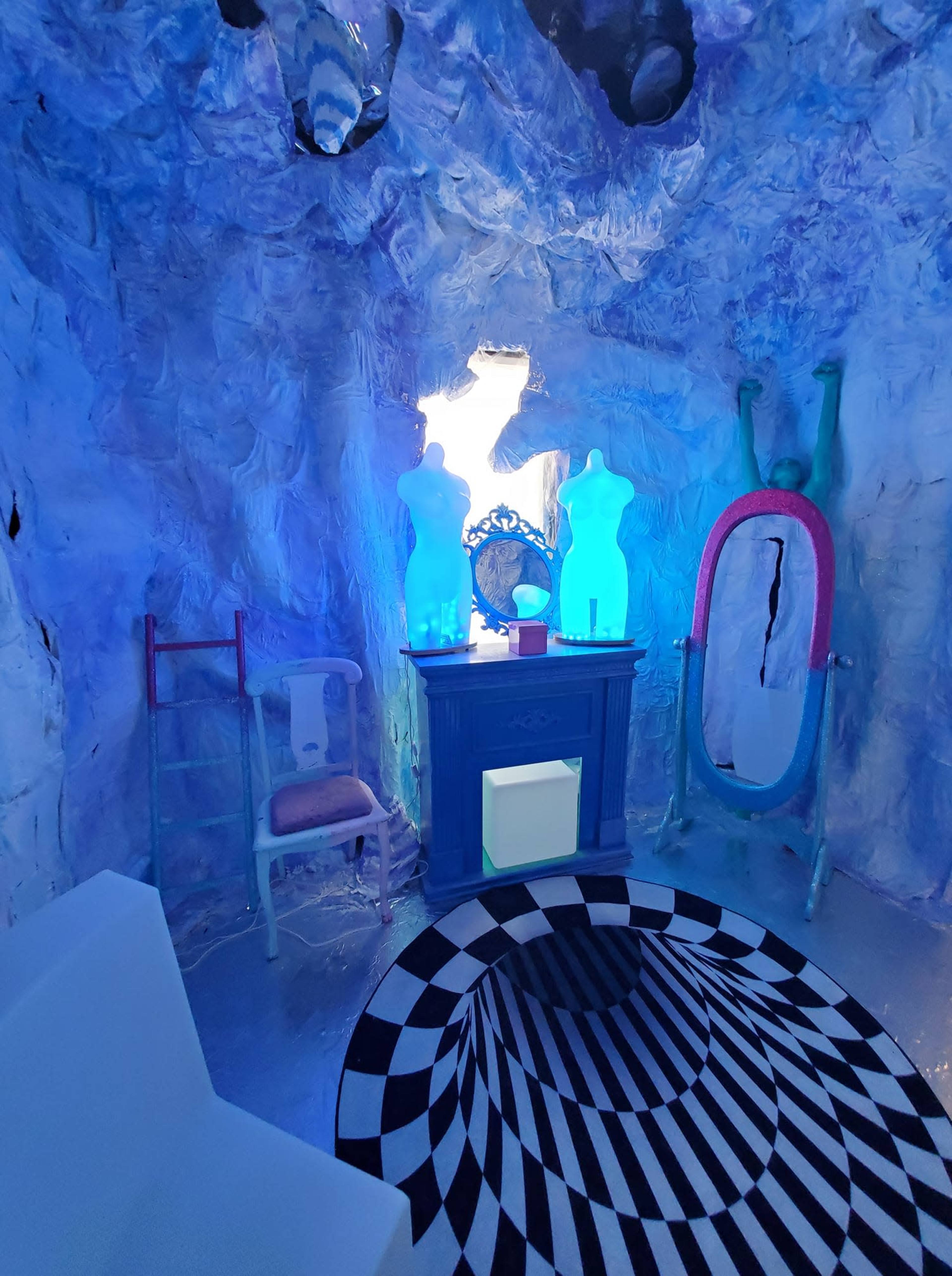 The image shows a vivid, abstract interior space featuring illuminated mannequins, a large oval mirror, and a checkered rug.