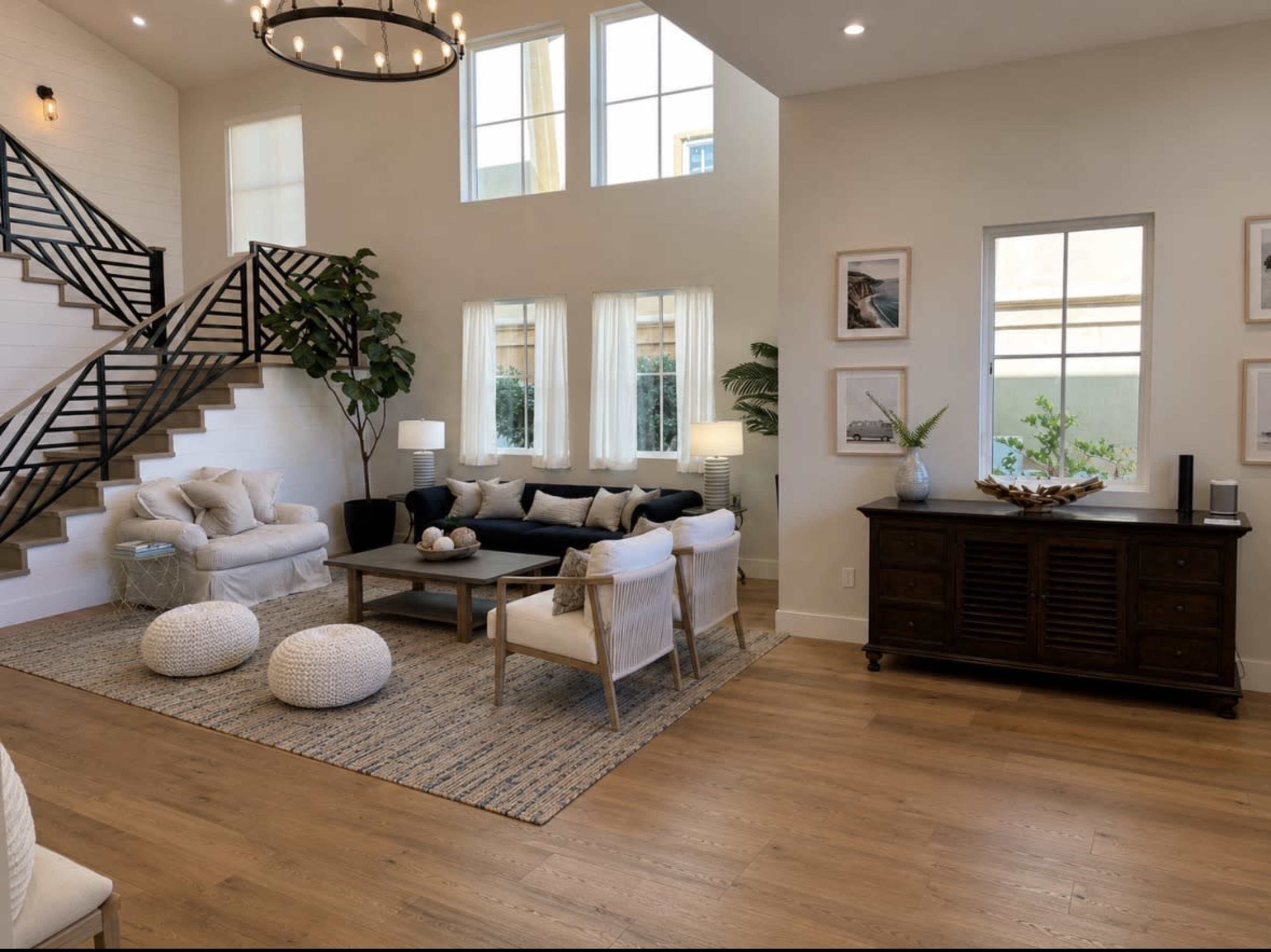 The image shows a modern living room with a large sectional sofa, a coffee table, and decorative elements, featuring a staircase in the background and ample natural light from large windows.