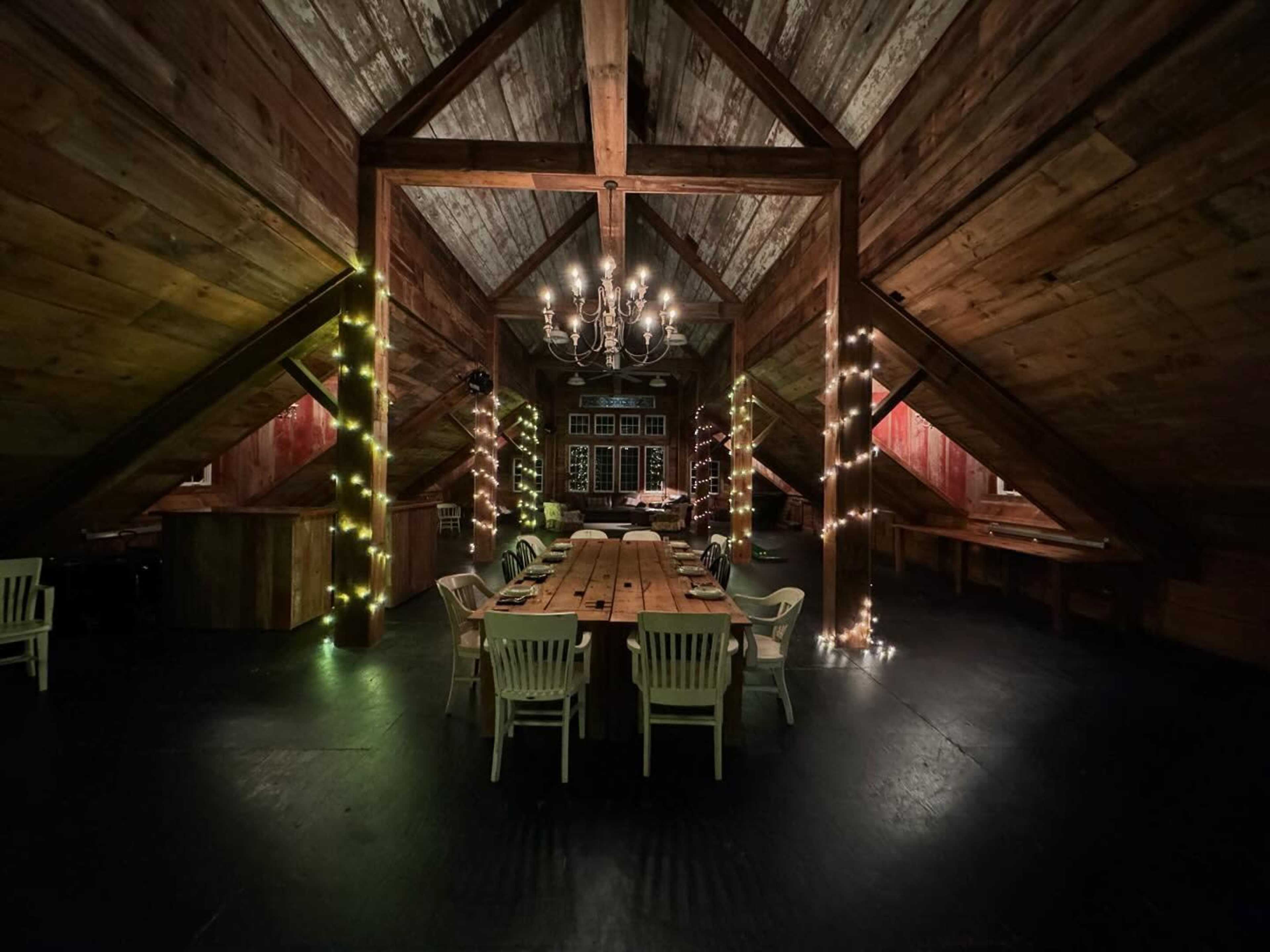 The image shows a rustic dining room with wooden beams, a long table, and soft lighting from string lights along the walls.