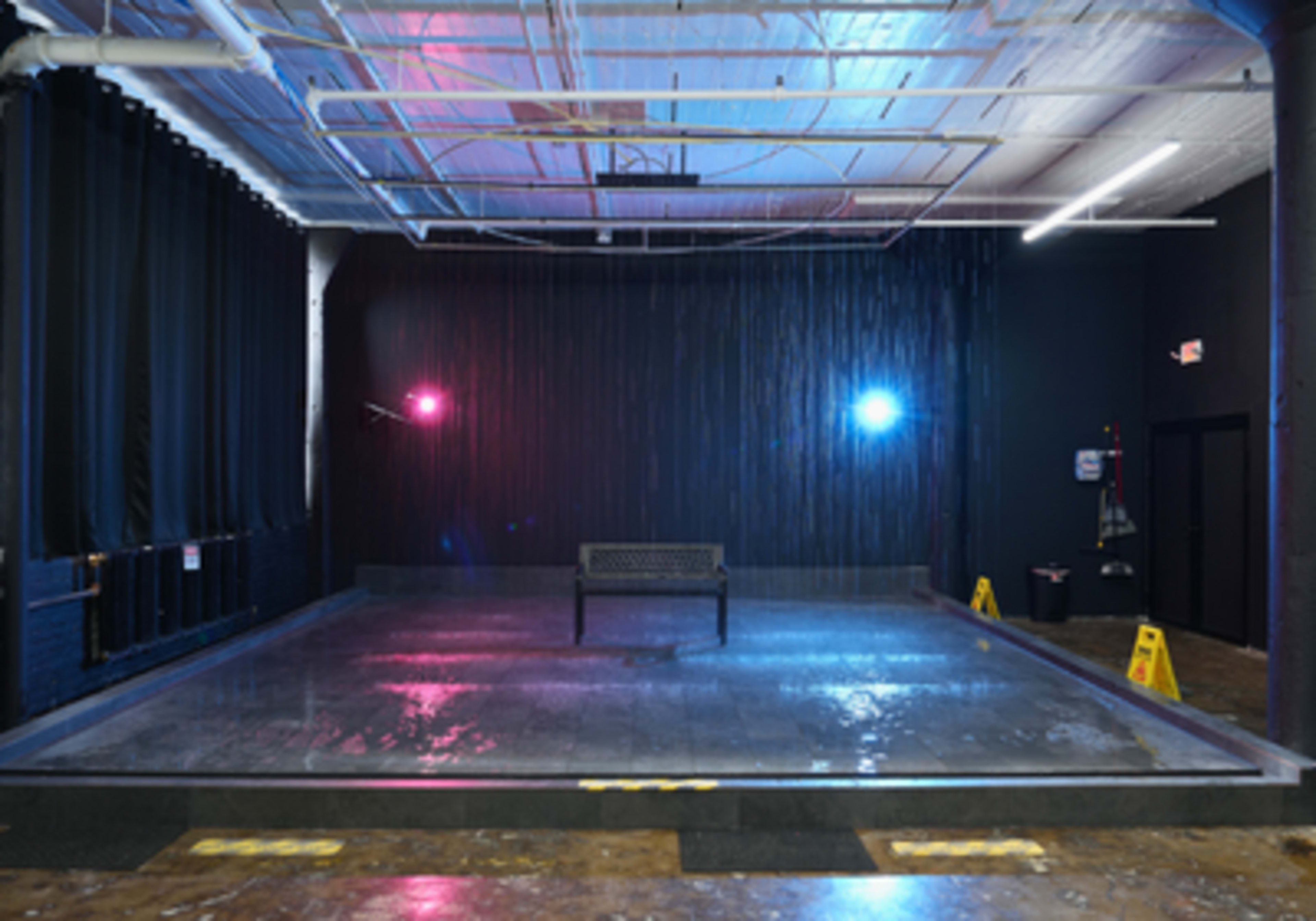 The image shows a dark stage area with a bench in the center, illuminated by blue and pink lights, and featuring a water cascade in the background.