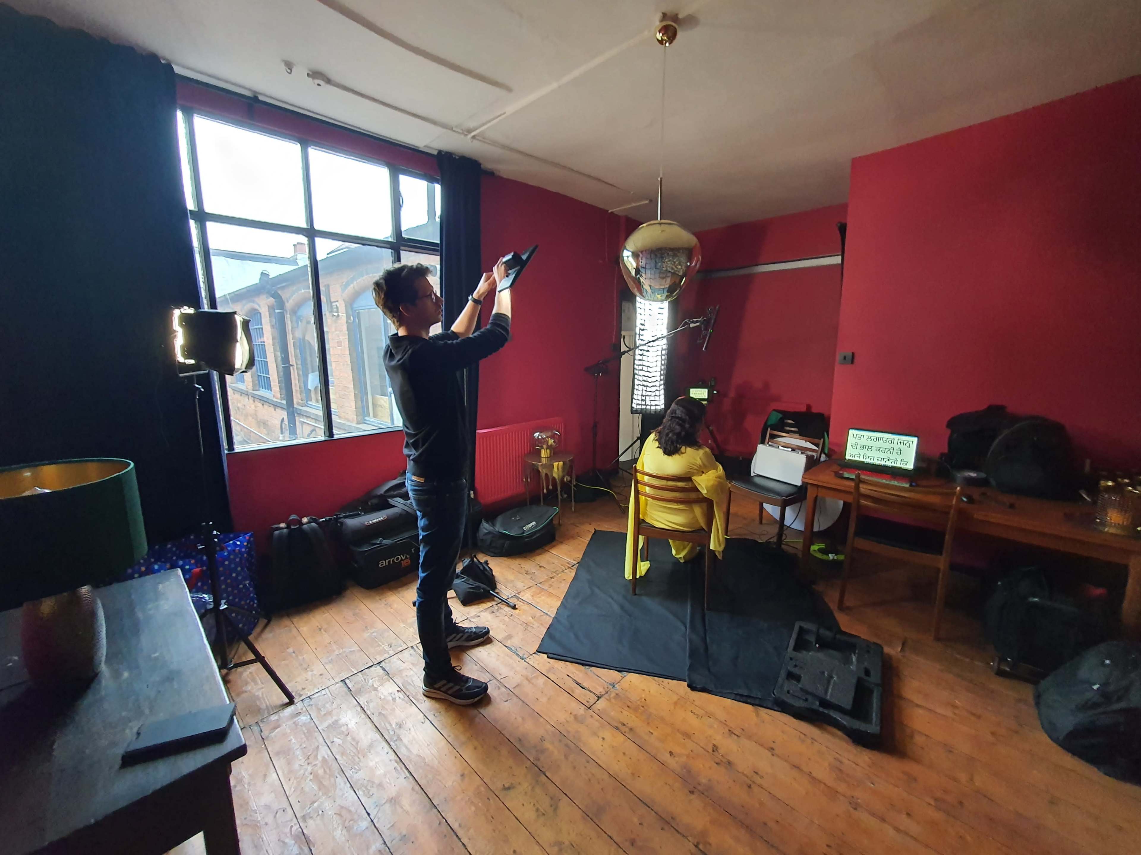 A person is setting up a camera to photograph a model sitting on a chair in a room with red walls and a disco ball hanging from the ceiling.