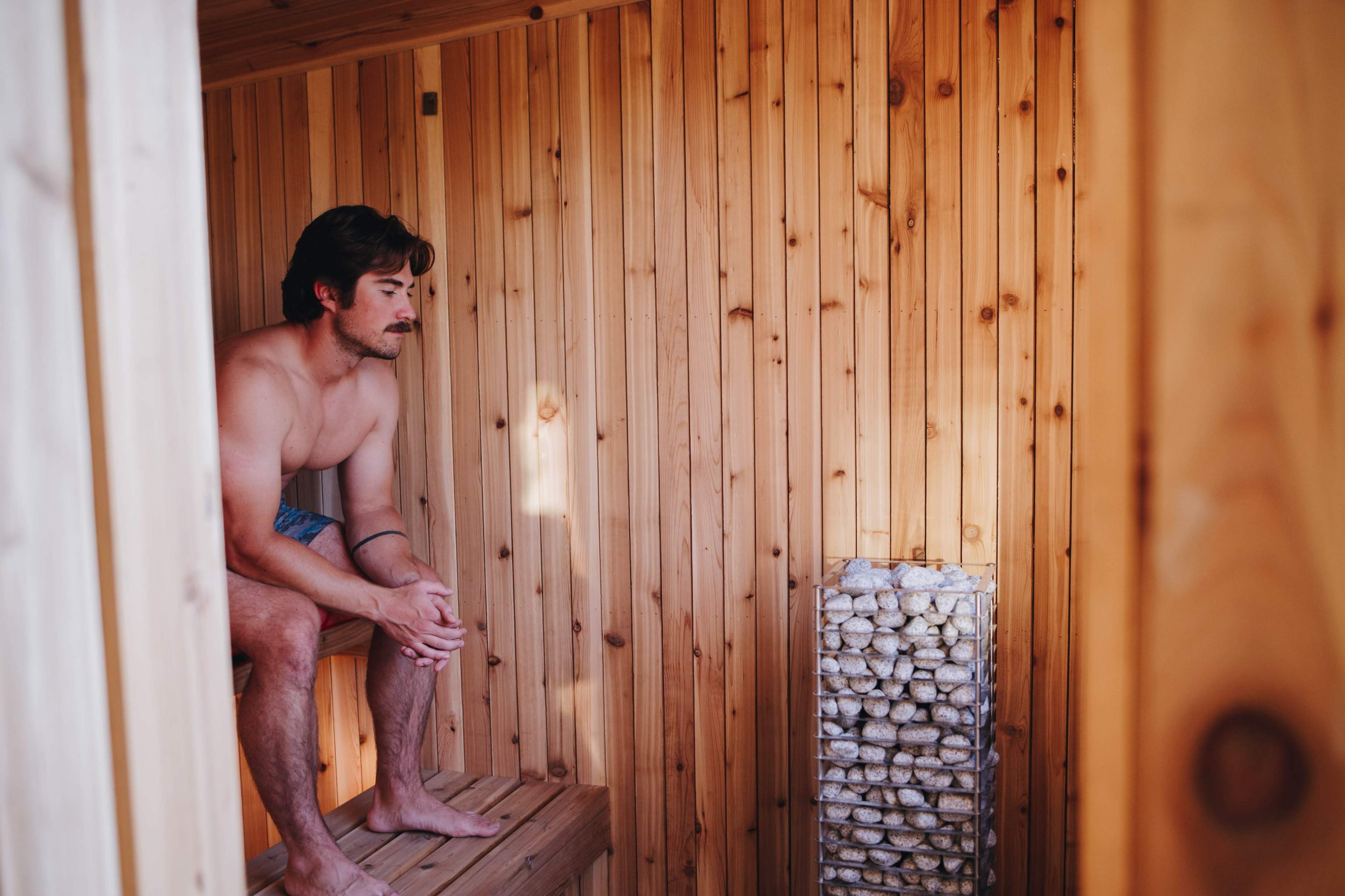 A man sits on a wooden bench inside a wooden sauna, with a stone heater nearby.