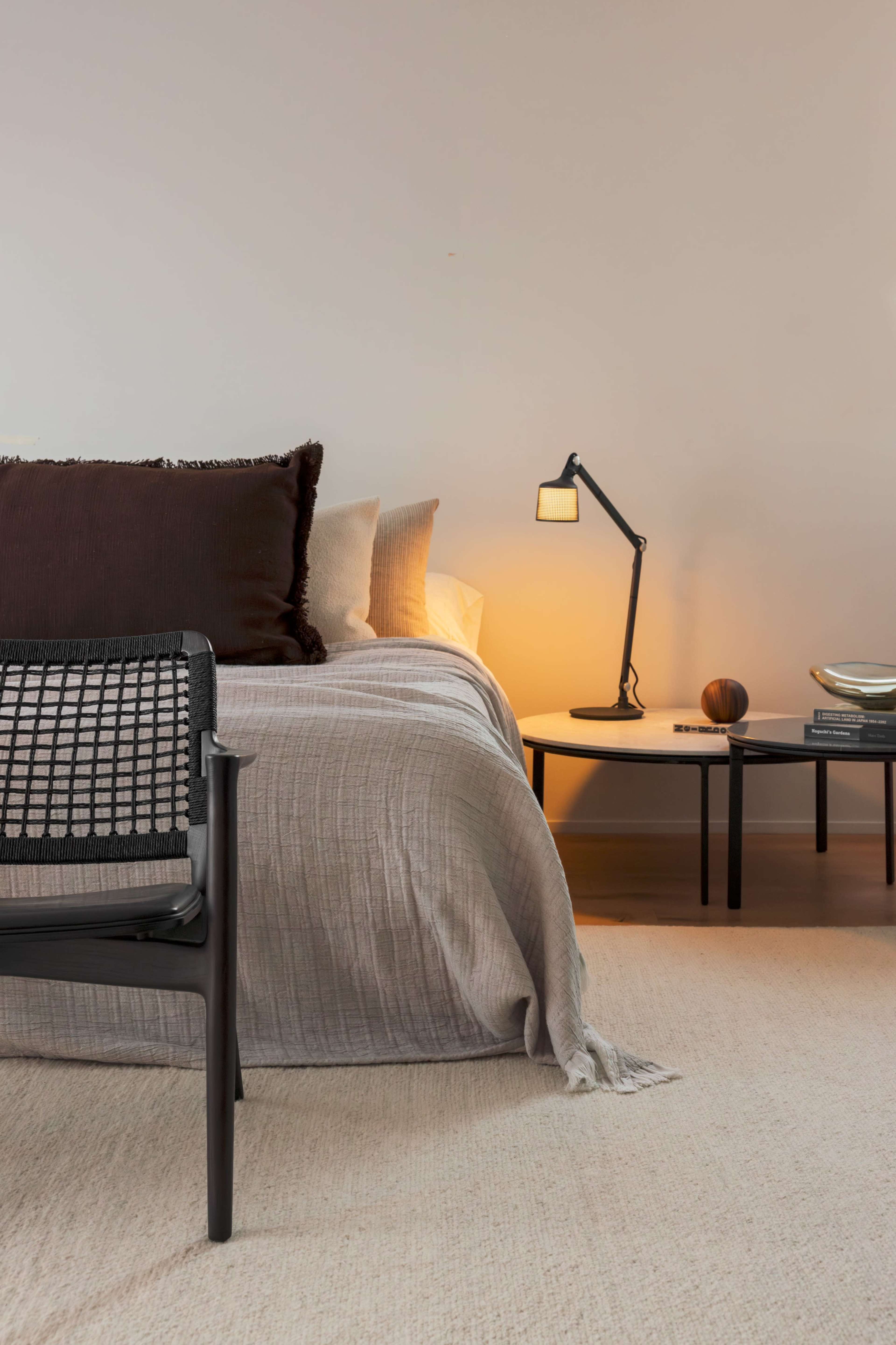 The image shows a modern bedroom featuring a bed with neutral-toned bedding, a minimalist side table, and a chair with a woven backrest.